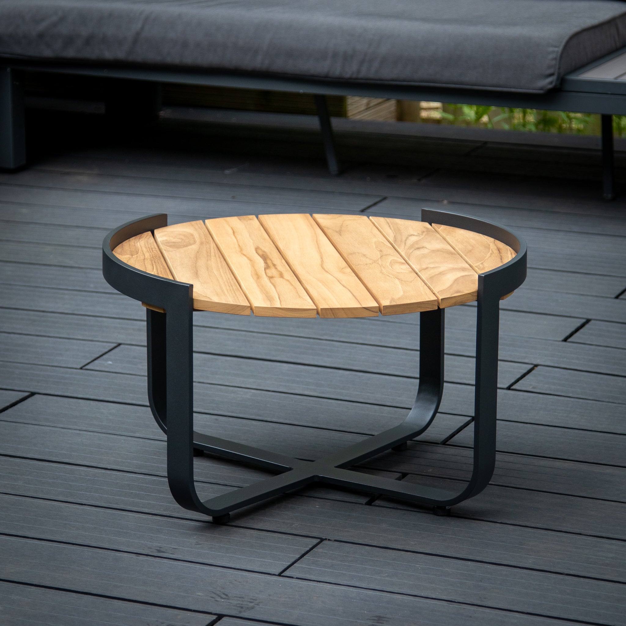 Bloom Coffee Table with Teak Top in Charcoal featuring a black metal frame on a dark wooden deck, complemented by a grey cushioned bench in the background. Perfect for outdoor furniture settings.