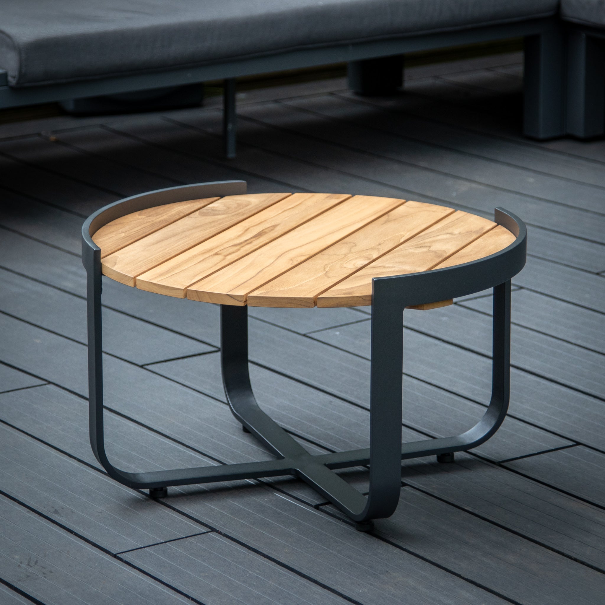 The Bloom Coffee Table with Teak Top in Charcoal features a round wooden teak top and a black metal frame, perfectly complementing the gray deck and outdoor seating in the background. This piece of outdoor furniture effortlessly combines elegance and durability for your patio space.