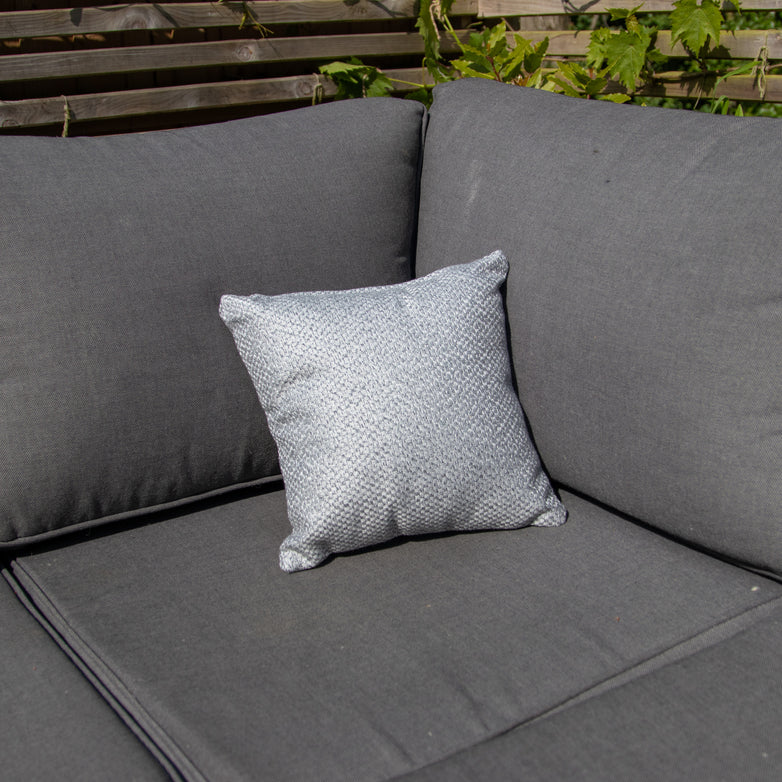 An Agora Senda Ceniza Small Scatter Cushion - 25m x 25cm, made from solution-dyed acrylic, is placed on a gray outdoor sofa with a wooden fence and greenery in the background.