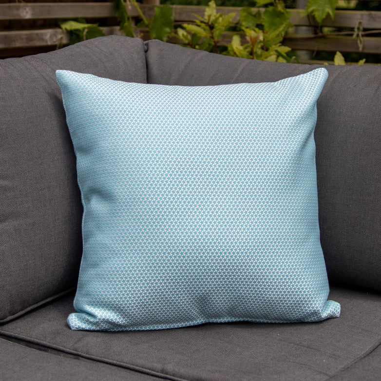 An Agora Scala Celeste Medium Scatter Cushion (45cm x 45cm) in light blue sits elegantly on a dark gray outdoor sofa, with greenery in the background, offering a luxurious touch to your outdoor setting.