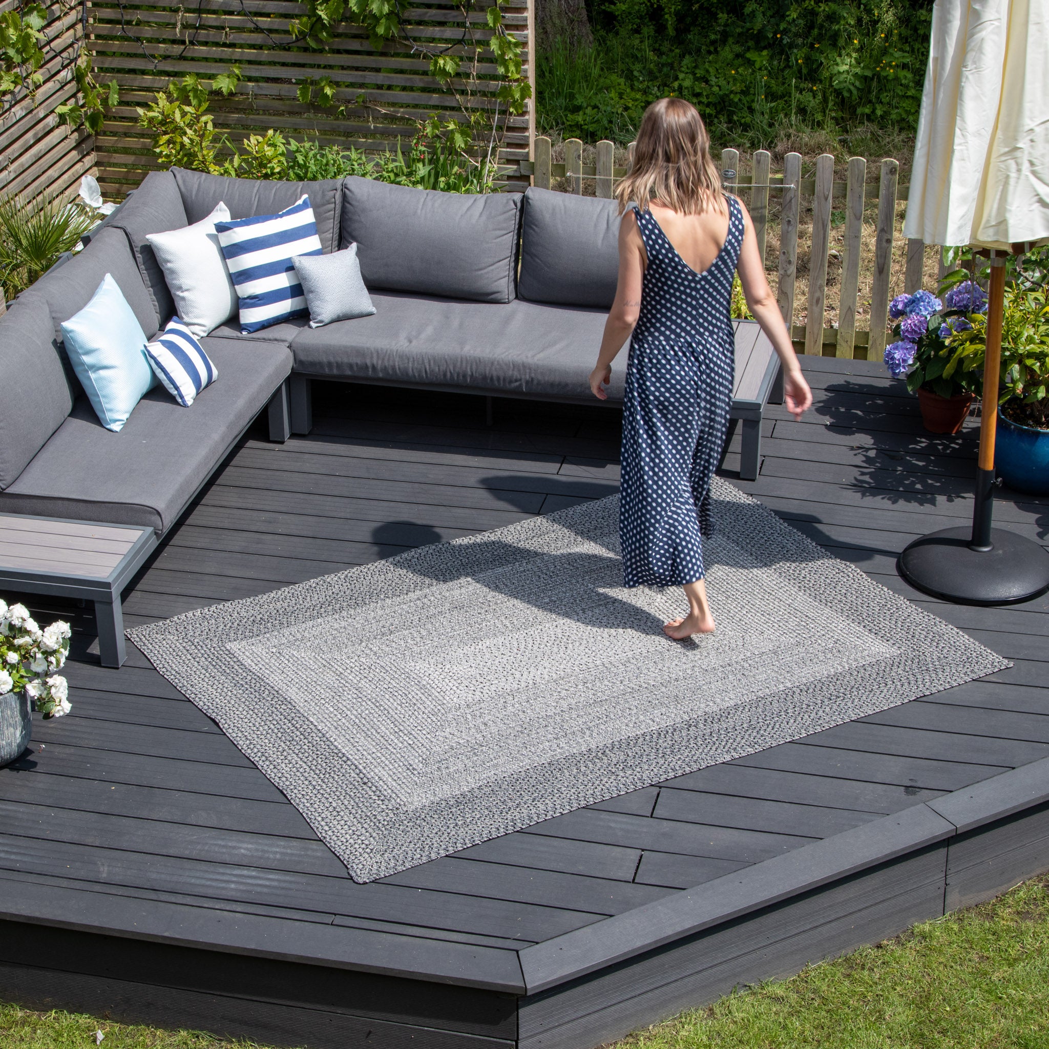 A woman in a blue polka dot dress strolls on an Artemis 366cm x 274cm Weatherproof Indoor and Outdoor Rug in Slate Grey in a backyard patio adorned with gray outdoor furniture and pillows.