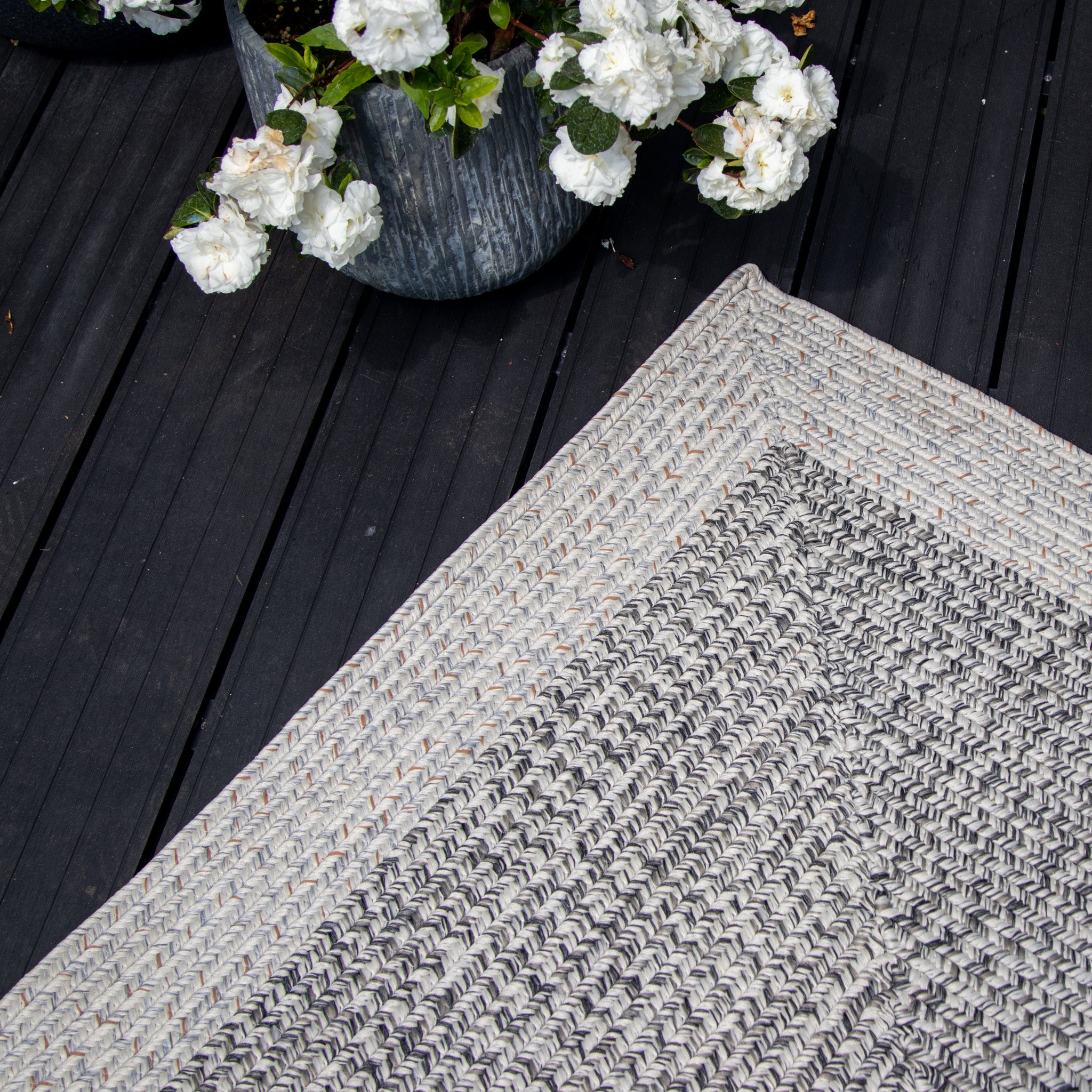 A Hades 290cm x 190cm Weatherproof Indoor and Outdoor Rug in Cobble Grey sits gracefully on a dark wooden deck, accompanied by two pots of white flowers.