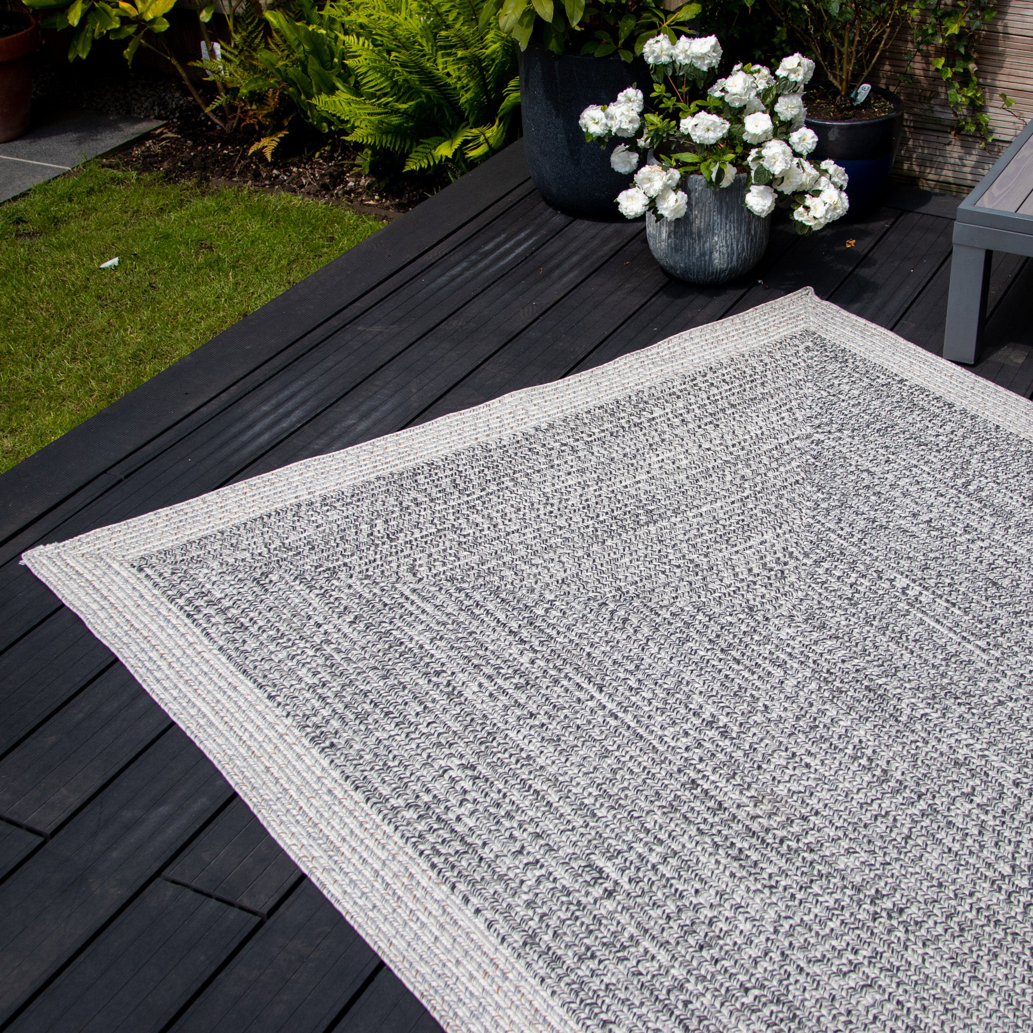 A Hades 290cm x 190cm Weatherproof Indoor and Outdoor Rug in Cobble Grey on a dark wood deck, decorated with potted white flowers in the background.