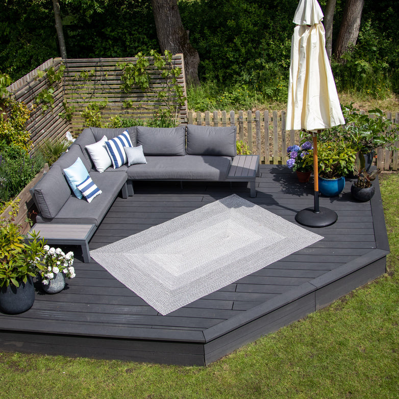 Outdoor patio with a UV resistant Walnut Grey sectional sofa, blue and white cushions, a Hestia 220cm x 160cm Weatherproof Indoor and Outdoor Rug in Walnut Grey, potted plants, and an umbrella on the wooden deck.