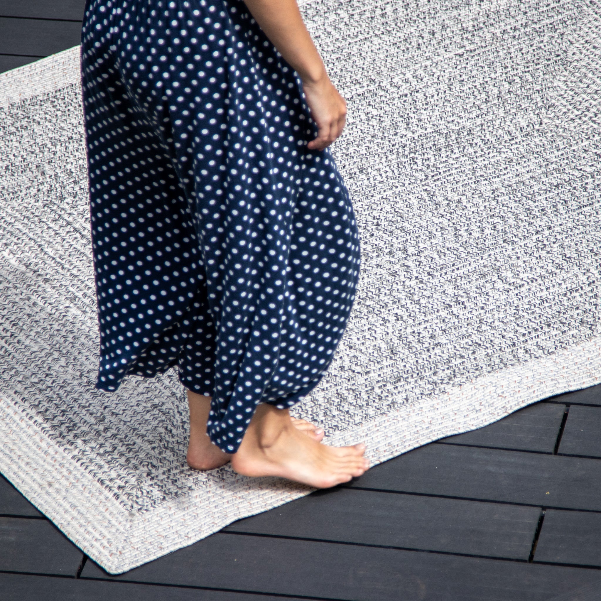 A person in a blue polka dot dress walks barefoot on a Hades 290cm x 190cm Weatherproof Indoor and Outdoor Rug in Cobble Grey over dark wooden flooring.