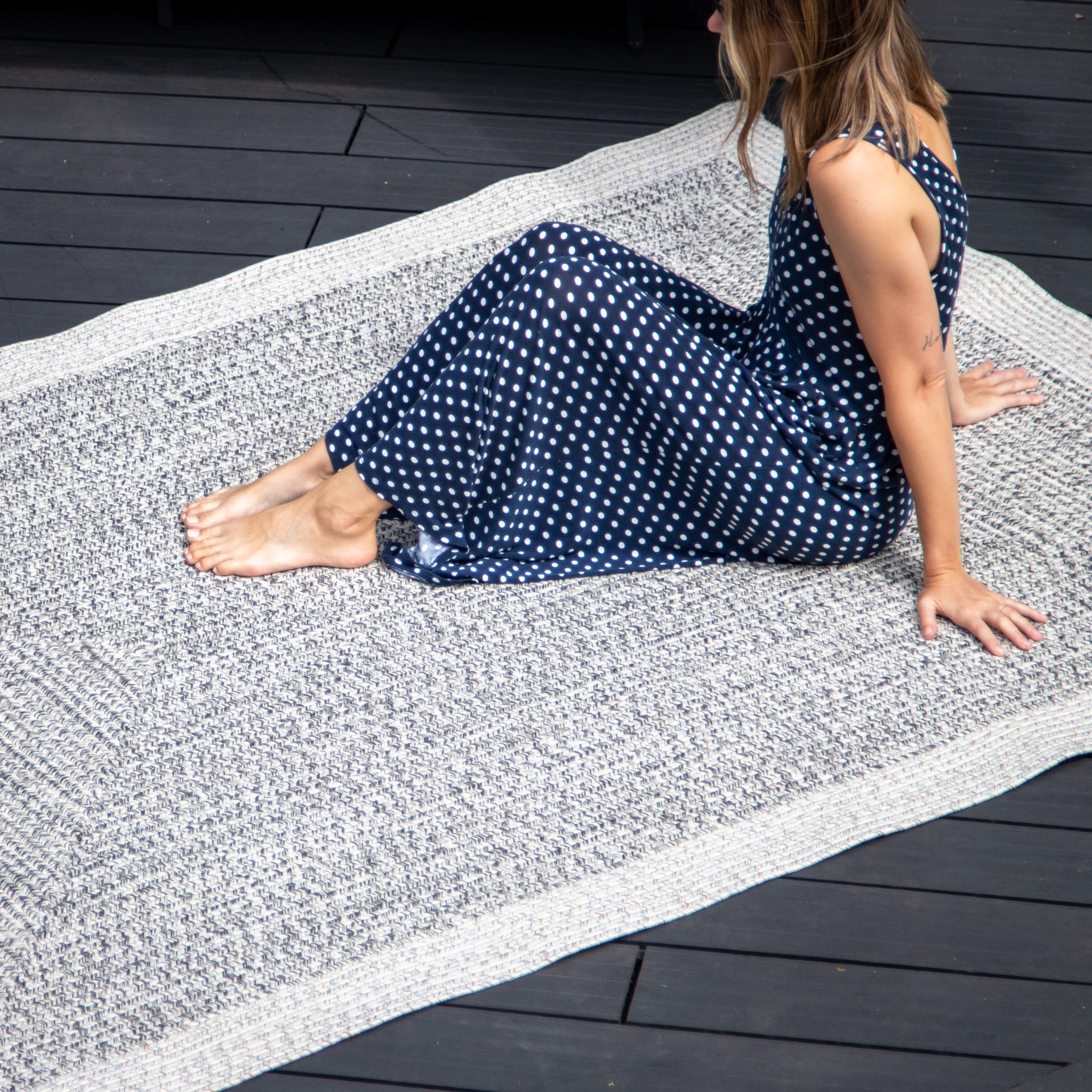 Person in a navy polka-dot dress sitting on the Hades 290cm x 190cm Weatherproof Indoor and Outdoor Rug in Cobble Grey, featuring a stylish grey and white design, on a dark wooden floor.