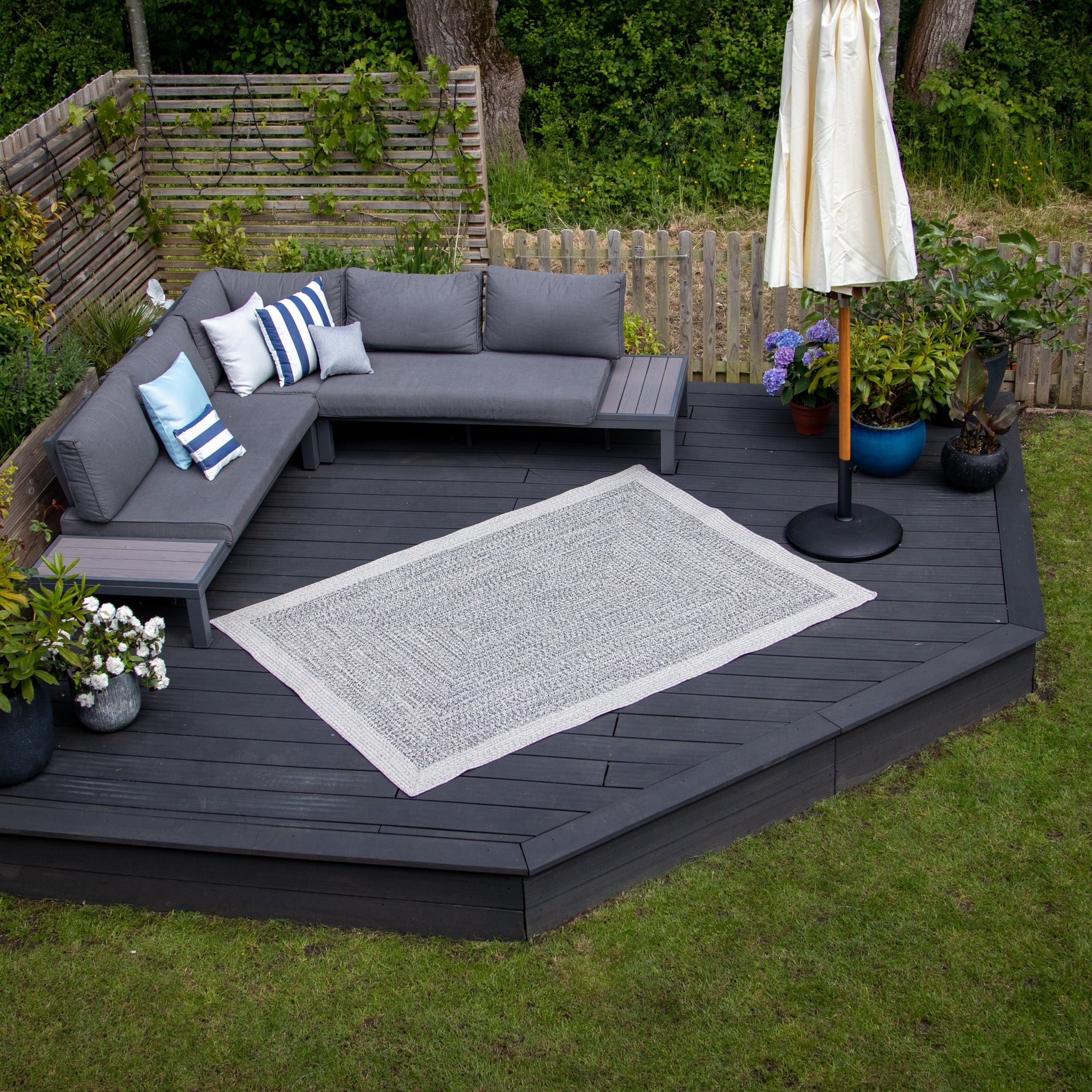 Outdoor patio with a UV-resistant Cobble Grey sectional sofa, white and blue pillows, umbrella, potted plants, and a Hades 290cm x 190cm Weatherproof Indoor and Outdoor Rug in Cobble Grey on the wooden deck.
