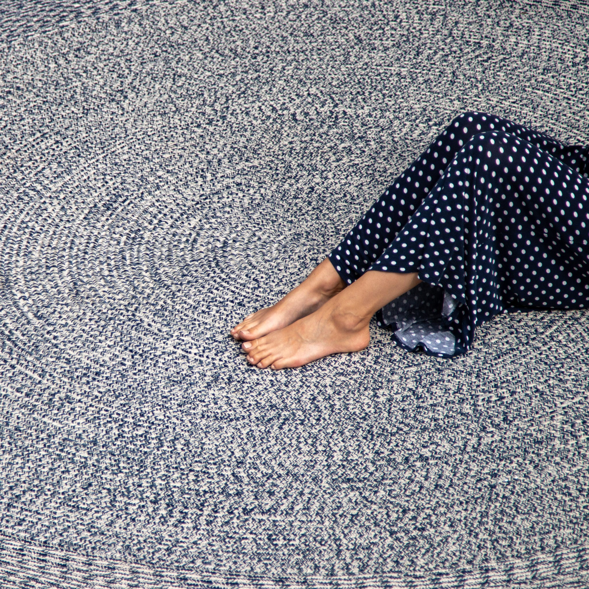 Bare feet and legs of a person in a polka-dot dress resting on an Apollo 300cm Round Indoor and Outdoor Rug in Prussian Blue with a textured circular pattern.