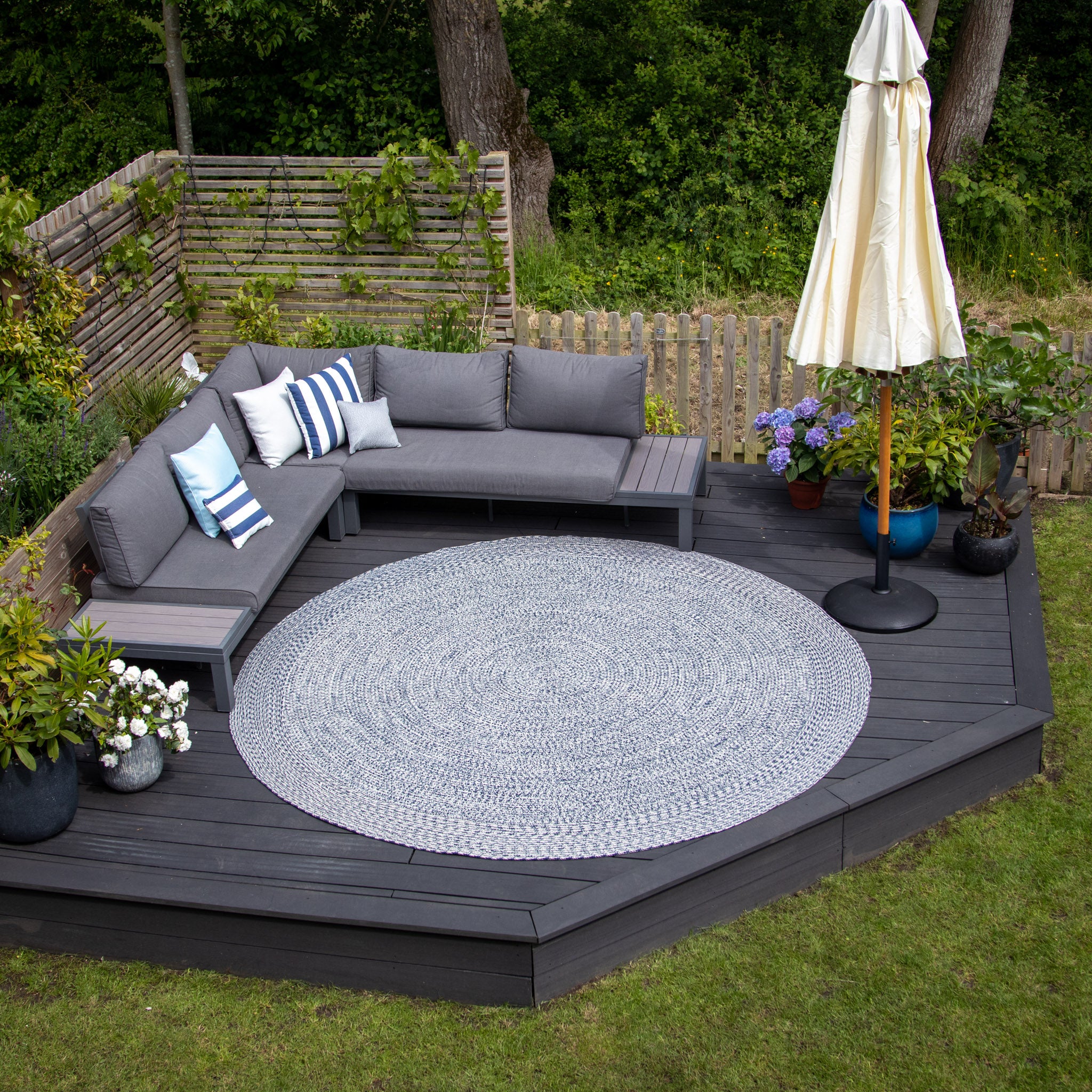 Outdoor patio with a gray sectional sofa, Apollo 300cm Round Indoor and Outdoor Rug in Prussian Blue, and umbrella, surrounded by plants and a wooden fence. The furniture is weatherproof and UV resistant for lasting beauty.