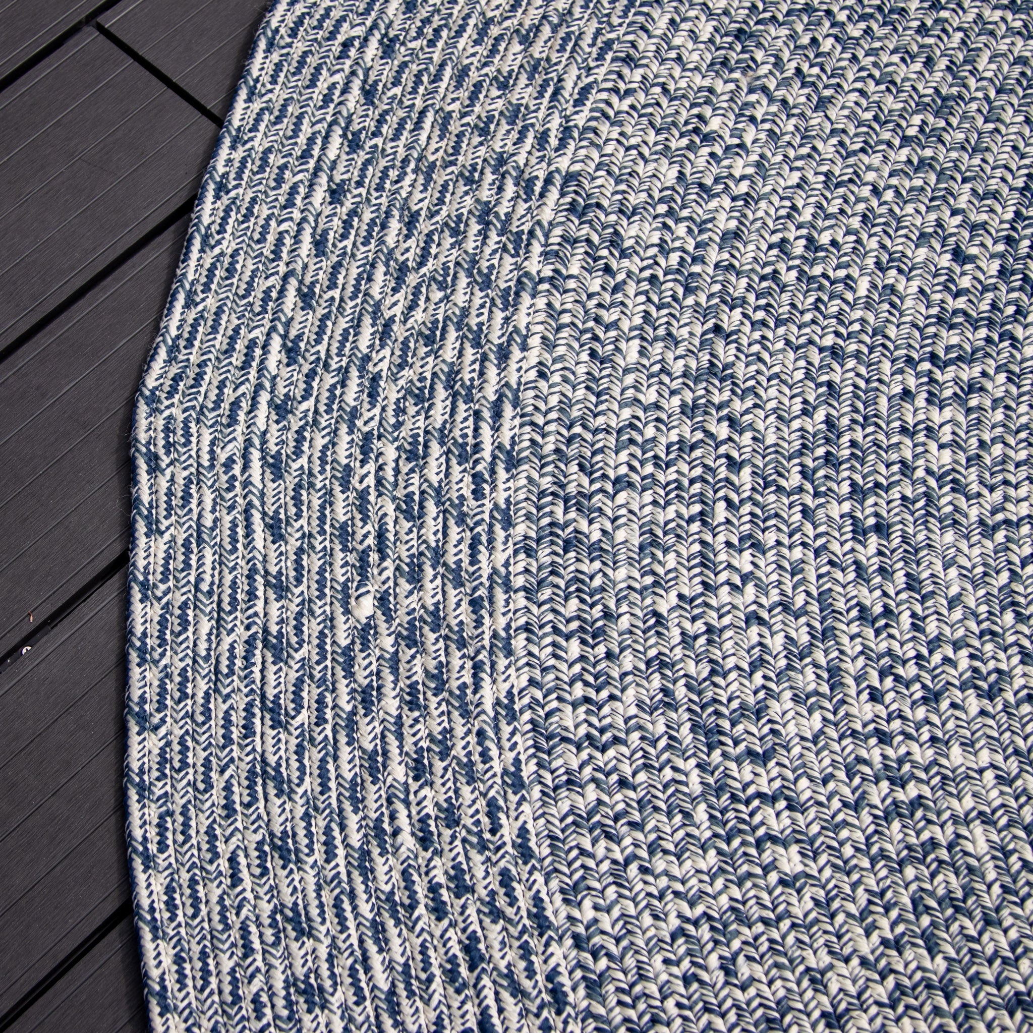 Close-up of the Apollo 300cm Round Indoor and Outdoor Rug in Prussian Blue, circular and woven in blue and white, partially covering a dark, textured floor. This weatherproof and UV resistant piece ensures durability in any setting.