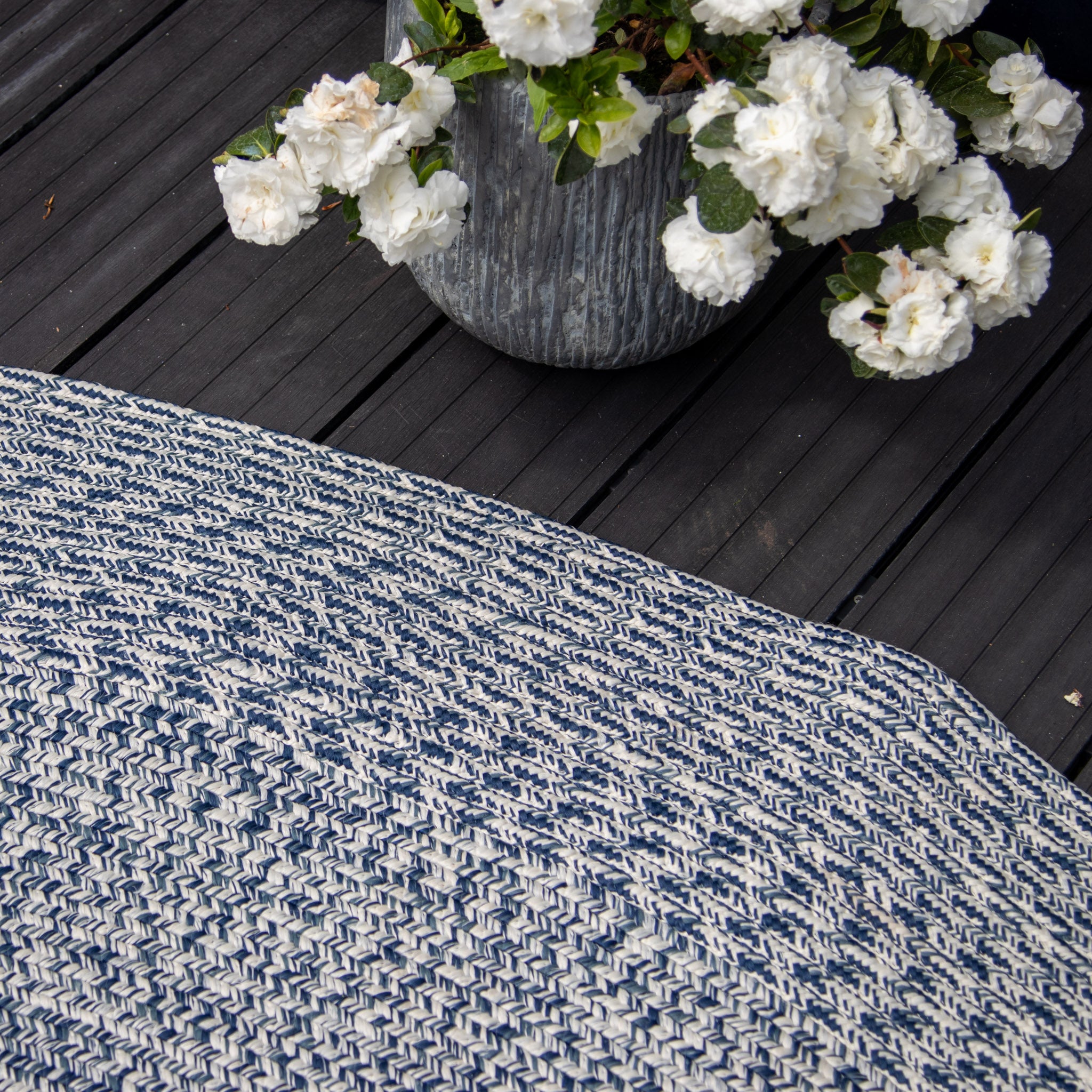 Close-up of a weatherproof Apollo 300cm Round Indoor and Outdoor Rug in Prussian Blue next to a large pot of white flowers on a dark wooden deck.