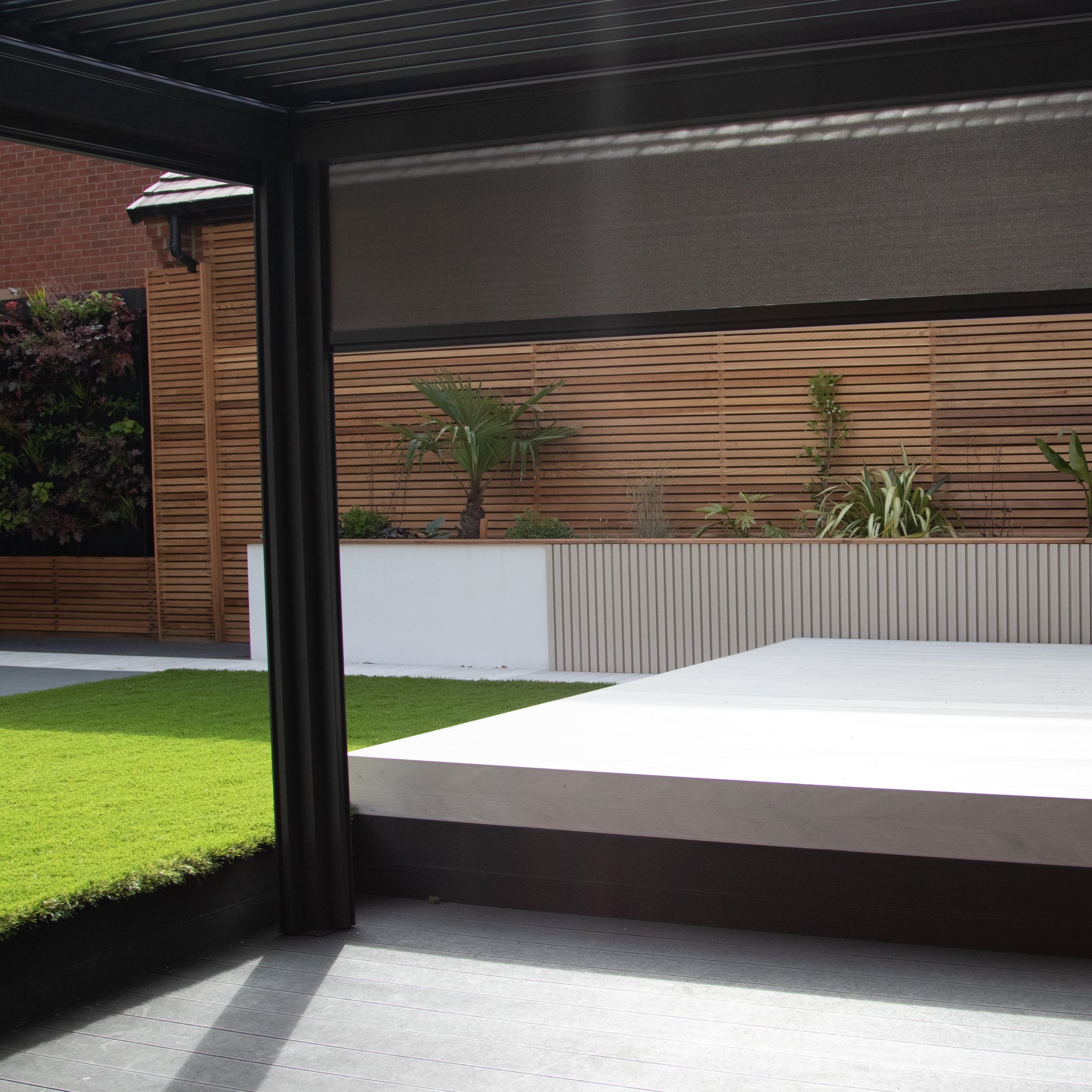 Modern patio with a wooden privacy fence, grassy area, and raised deck under a PergoSTET Plus 3m x 3m Square Pergola in Grey featuring louvred roof for adjustable sunlight, 3 drop sides, and LED lighting.