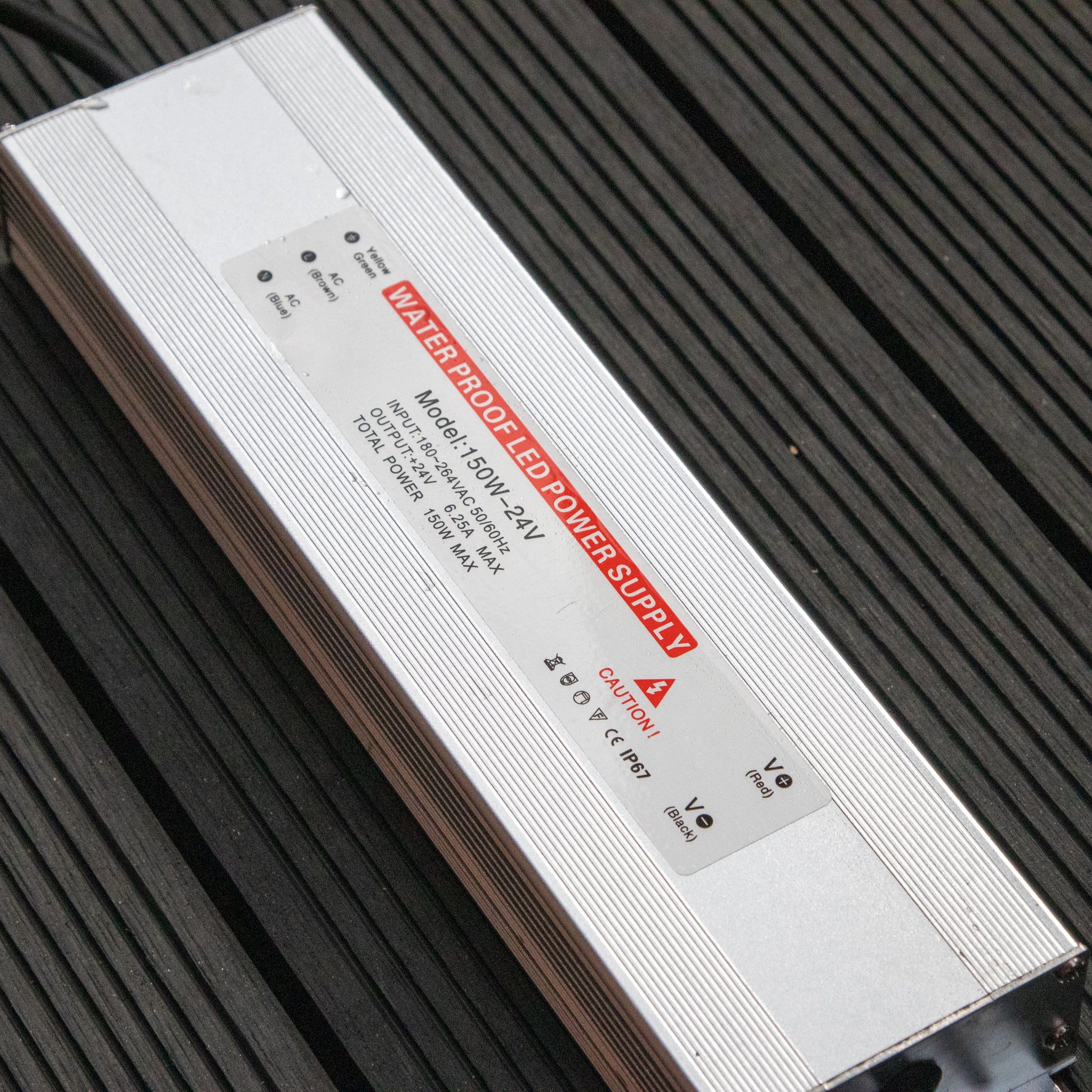 A rectangular waterproof power supply unit sits on a dark, textured surface, echoing the sleek design of modern pergolas. Labeled "Waterproofed Power Supply" in bold red, it complements outdoor innovations like the PergoSTET Plus 3m x 4m Rectangular Pergola with LED lighting in grey.