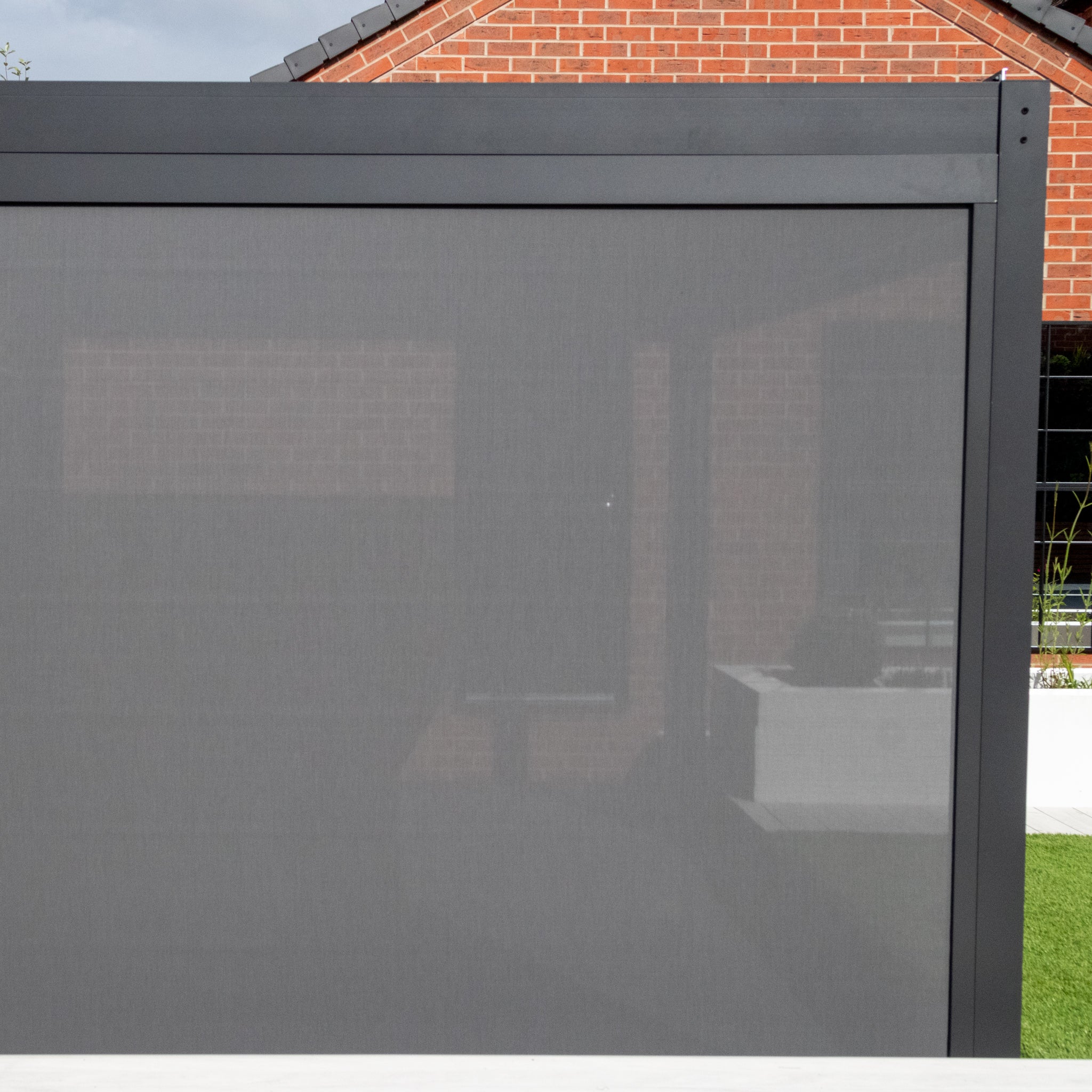 A dark gray privacy screen complements the brick house, forming a cozy outdoor area perfect for the stylish PergoSTET Plus 3m x 3m Square Pergola with 3 Drop Sides and LED Lighting in Grey.