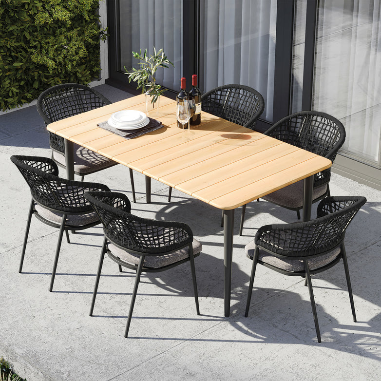 Kalama 6 Seat Rectangular Dining Set with a weatherproof aluminium frame, teak table in charcoal, black chairs, and place settings for six on a concrete patio near a glass door.