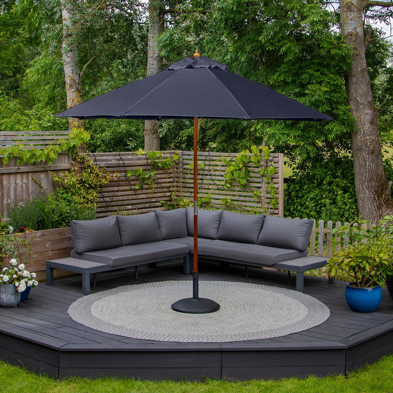 A corner patio with gray sofas, a Kratos 3m Round Wooden Parasol in Navy providing UV rating 50+ protection, a round rug, and greenery surrounding the wooden fenced area.