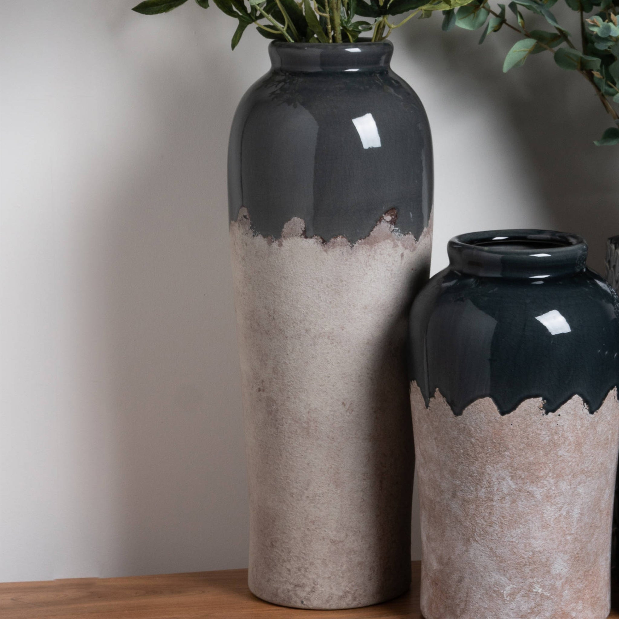 The Large Conran Vase, featuring a dual textured finish with a matte beige bottom and glossy dark gray top, is ideal for long stem floral arrangements and makes an elegant display on a wooden surface.