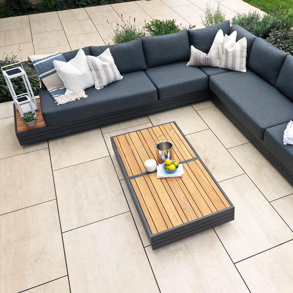 Outdoor patio with a gray sectional sofa, decorated with white pillows, surrounding a Panama Teak Coffee Table featuring a teak wooden top adorned with fruit and drinks.