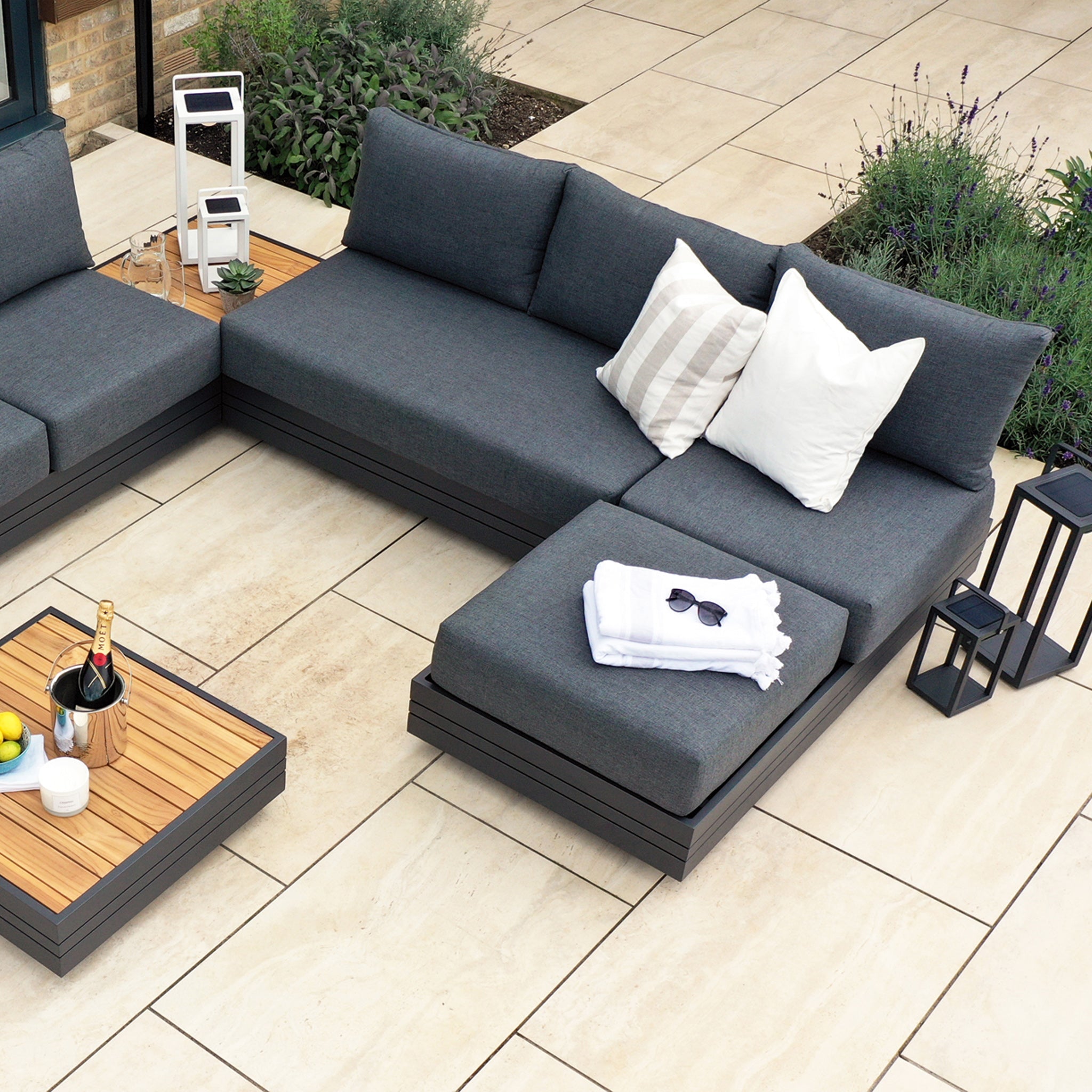 Modern patio with a Panama Corner Unit/Footstool in Charcoal, adorned with striped and white pillows, small table with drinks, and sunglasses on the footstool. Enjoy the versatility of this modular system designed for both comfort and style.