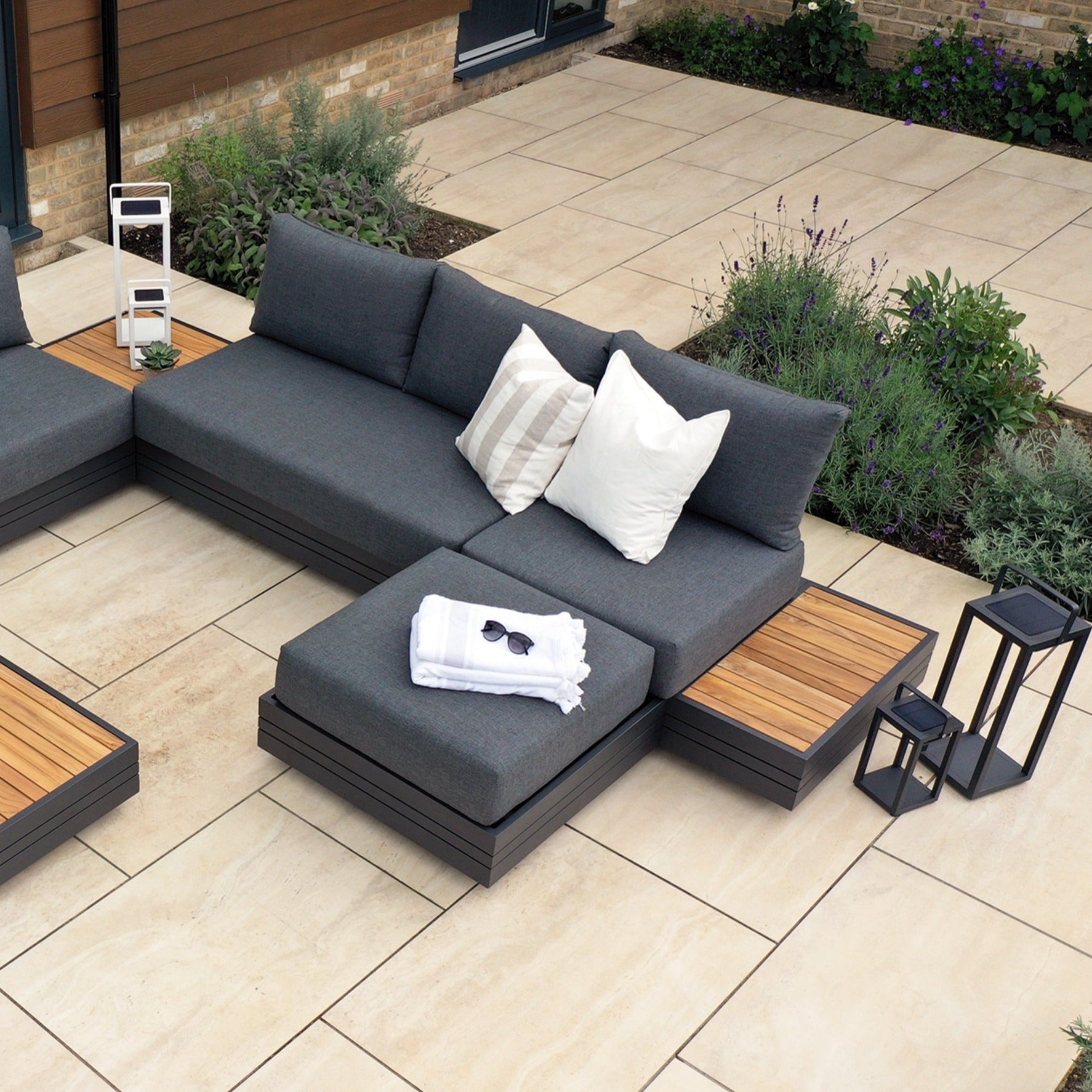 Contemporary outdoor patio seating area featuring Panama Corner Unit/Footstool in Charcoal with gray cushions, white pillows, and wooden side tables. The modular system includes the versatile corner unit and footstool. Nearby lanterns and green plants enhance the tranquil ambiance.