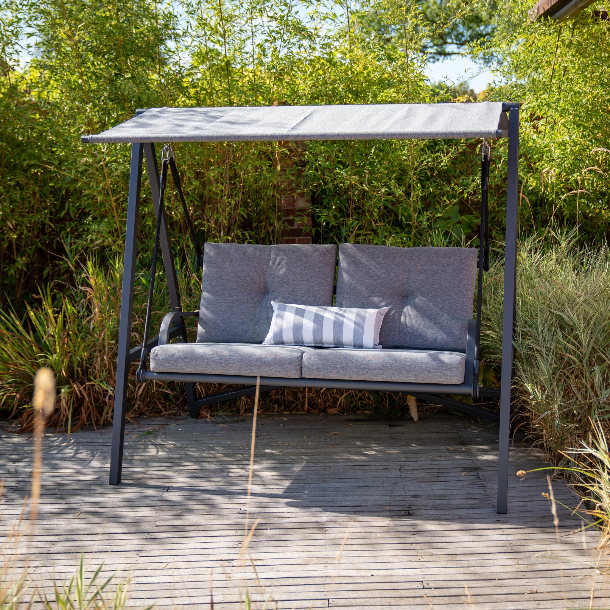 The Lima Aluminium Swing Seat in Washed Grey boasts a gray olefin canopy and cushions, accompanied by a stylish gray-and-white striped pillow. Surrounded by lush green plants, its powder-coated aluminium frame offers both durability and elegance for your outdoor sanctuary.