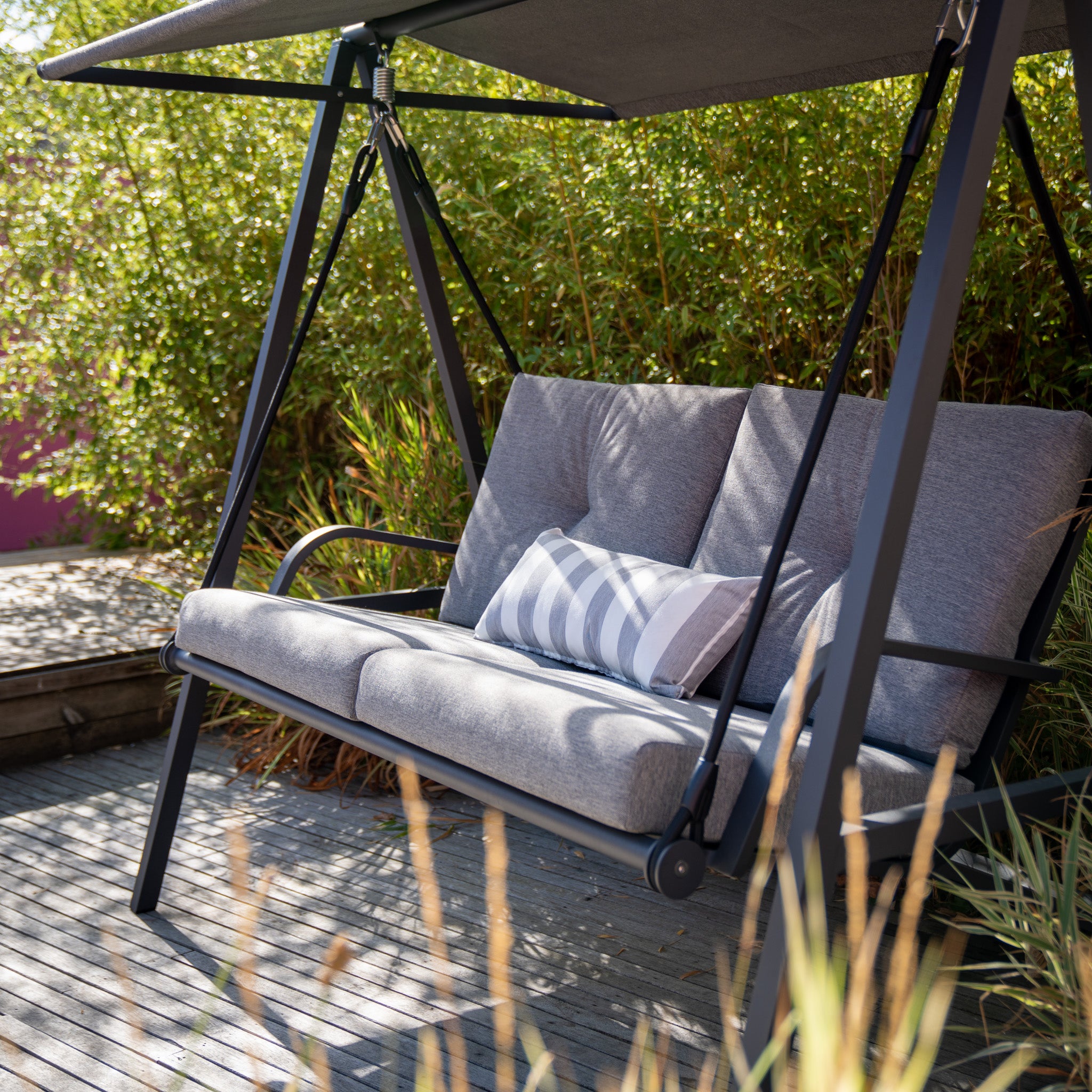 A Lima Aluminium Swing Seat in Washed Grey, complete with a striped pillow, sits on a wooden deck surrounded by tall greenery, featuring a powder-coated aluminium frame and an olefin canopy.