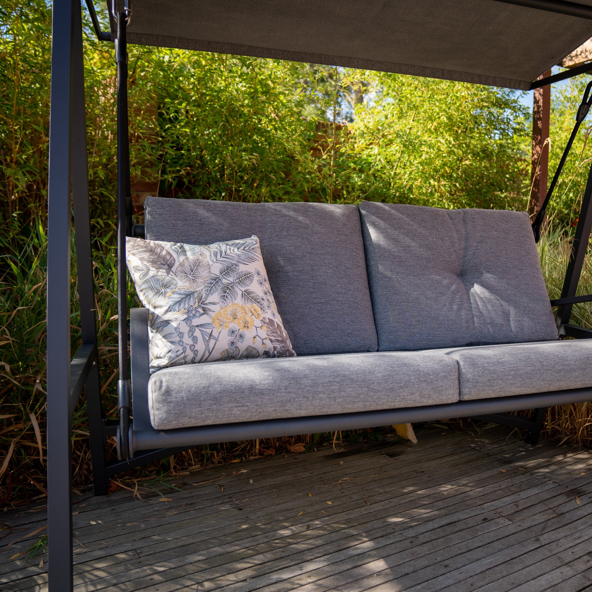 A Lima Aluminium Swing Seat in Washed Grey with a floral pillow sits on a wooden deck, surrounded by green foliage, featuring an olefin canopy and a powder-coated aluminium frame for added durability.