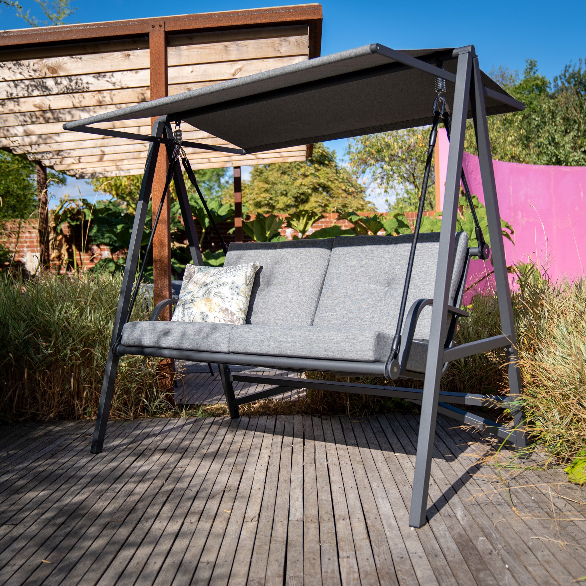 A Lima Aluminium Swing Seat in Washed Grey, adorned with an olefin canopy and a floral-patterned pillow, graces the wooden deck. It is surrounded by plants and trees and features a sleek powder-coated aluminium frame.