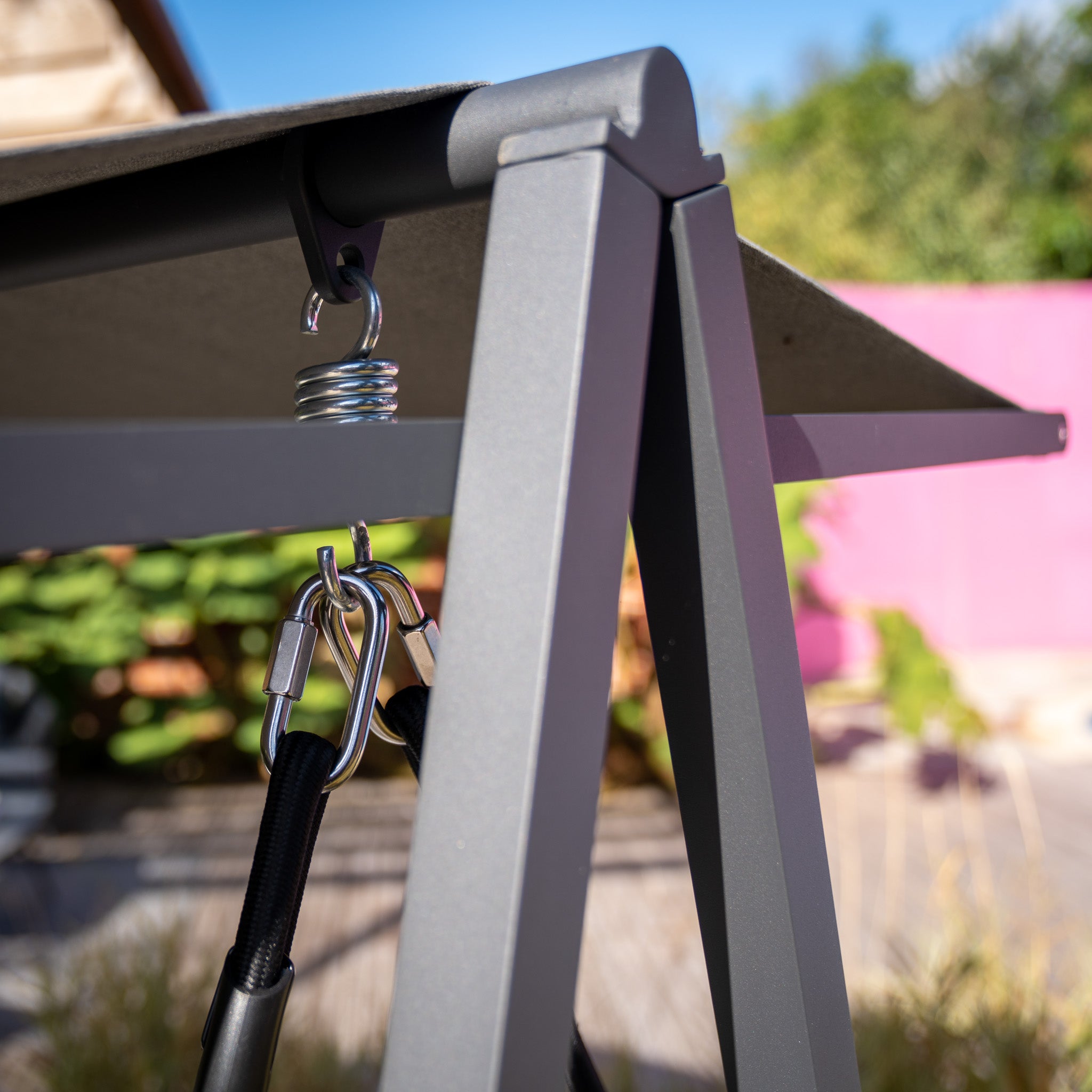 Close-up of the Lima Aluminium Swing Seat in Washed Grey with carabiner hooks and a spring attached to the powder-coated aluminium frame, featuring a blurred outdoor background.