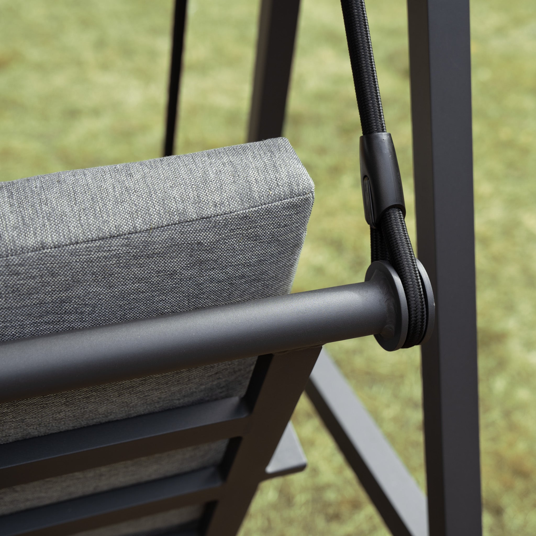 Close-up of a Lima Aluminium Swing Seat in Washed Grey with a gray cushioned seat and black ropes, set against a blurry green background. The powder-coated aluminium frame ensures durability, while an olefin canopy offers excellent weather resistance.