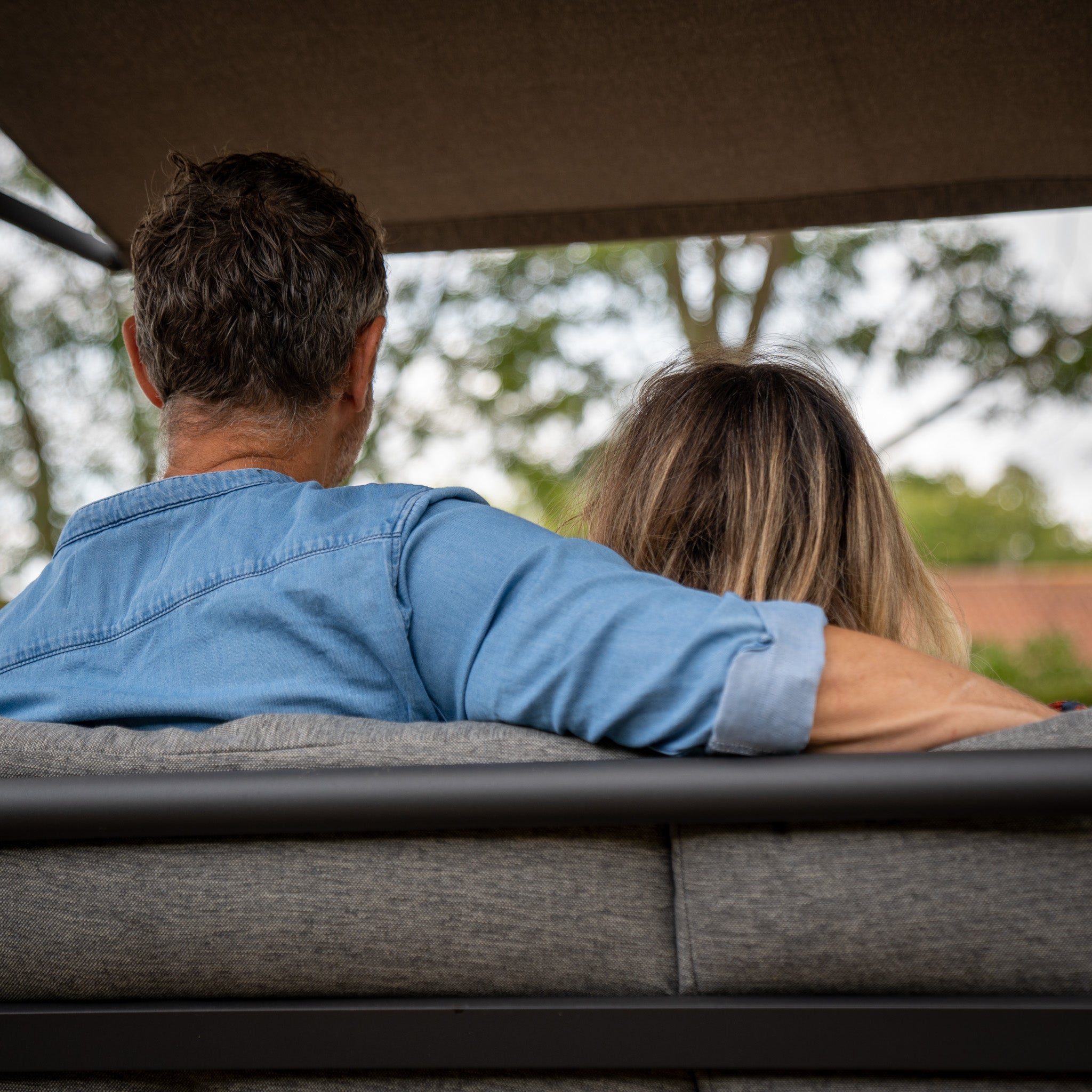 Two people sit closely on a Lima Aluminium Swing Seat in Washed Grey with an olefin canopy, nestled among greenery in the background, viewed from behind.