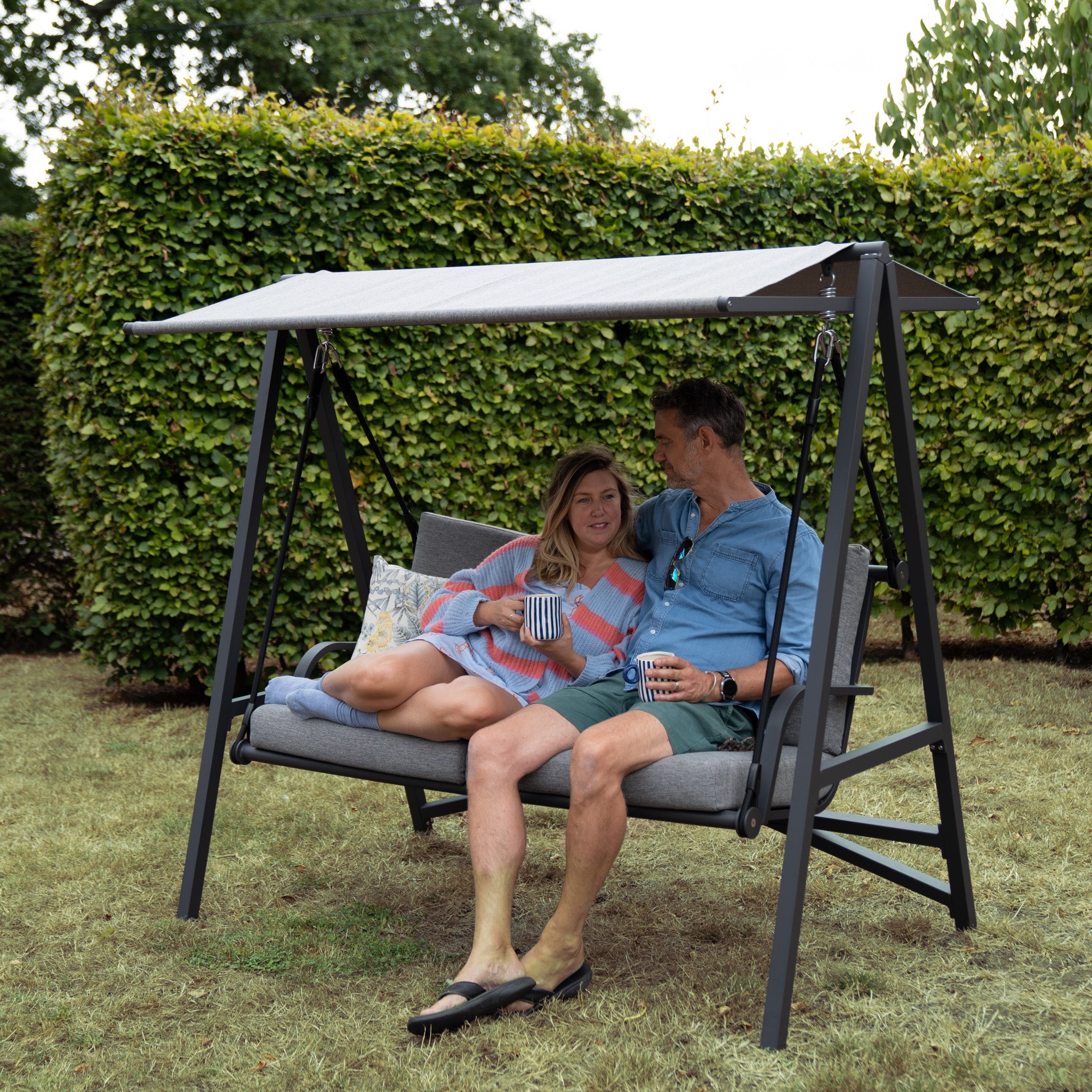 A couple sits on their Lima Aluminium Swing Seat in Washed Grey under an olefin canopy, smiling and holding mugs, with hedges and trees in the background.