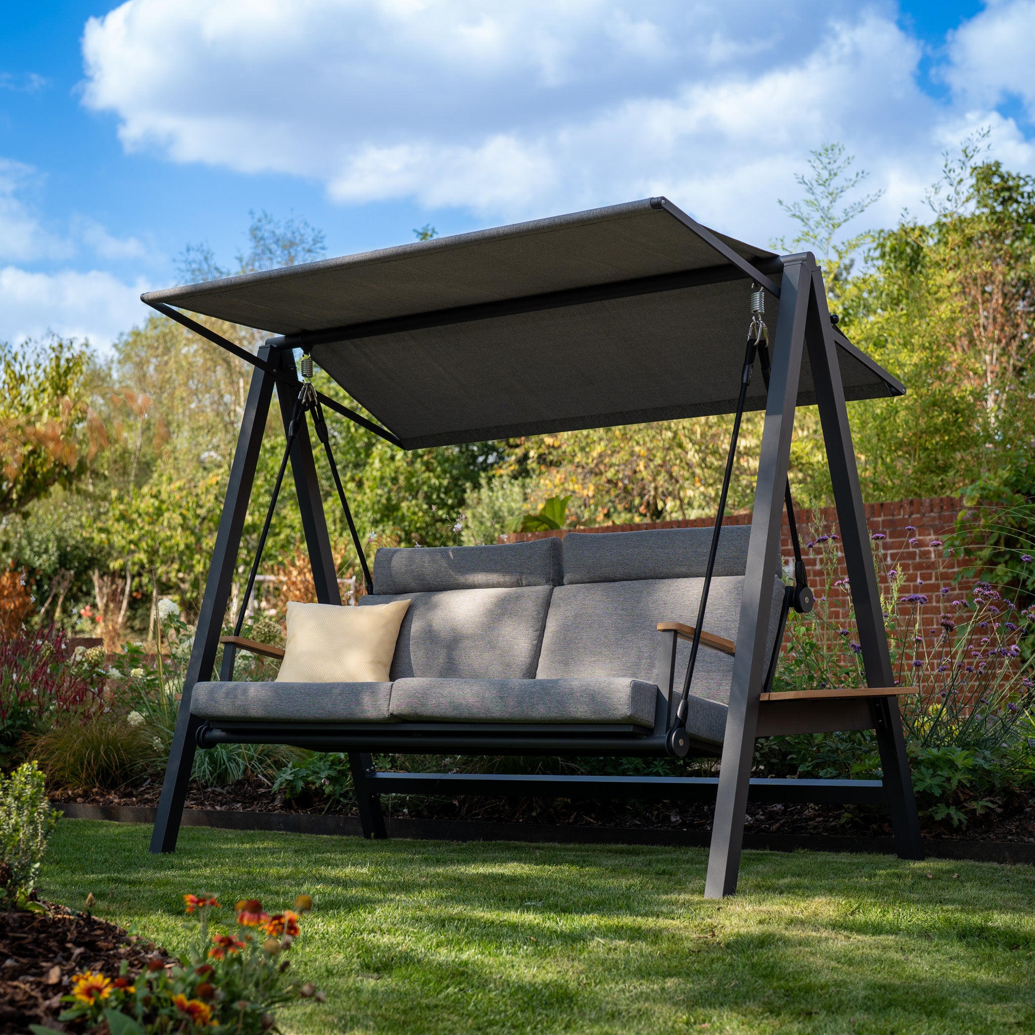 The Lima Deluxe Aluminium Swing Seat in Washed Grey, crafted with durable olefin material, features a canopy over a cushioned seat. Placed on a lawn adorned with flowers and trees in the background, it's perfect for enjoying sunny days.