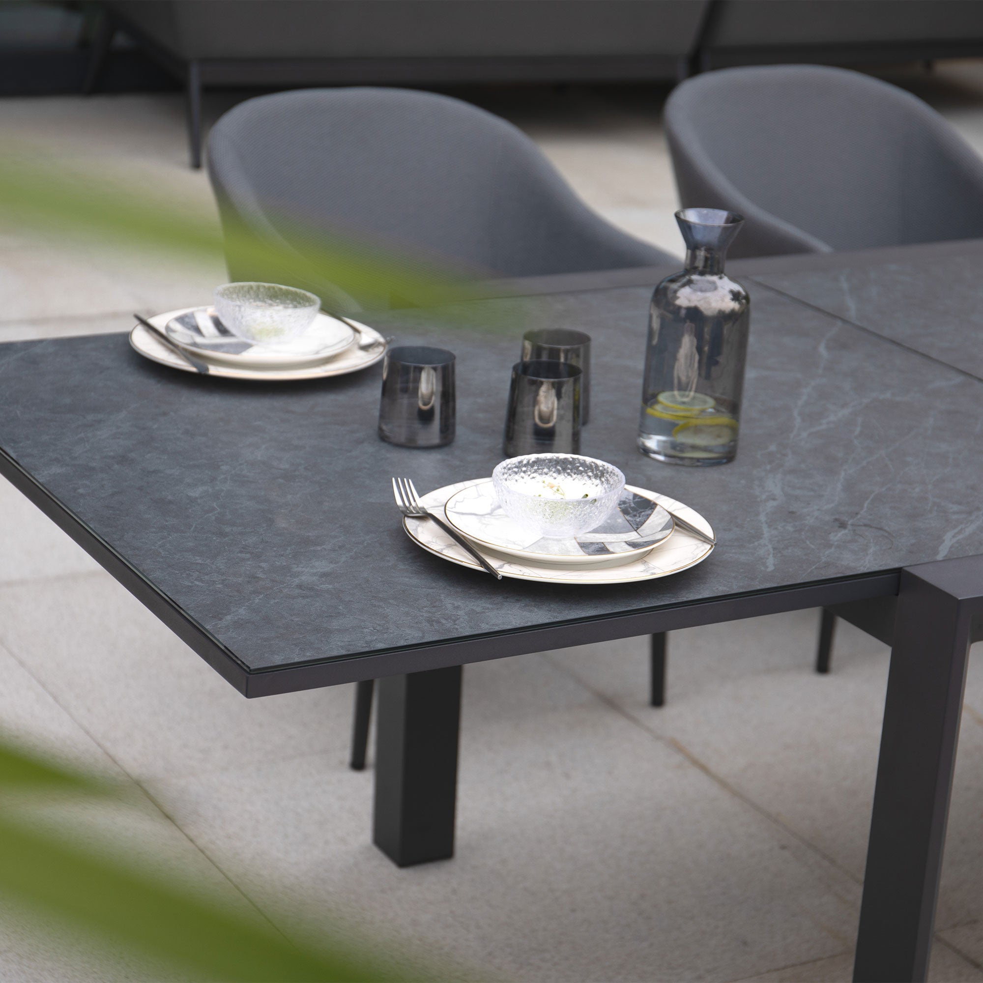 The Luna 8 Seat Outdoor Fabric Extending Ceramic Dining Set in Grey features modern stain-proof design, paired with sleek gray chairs, complemented by plates, glasses, and a carafe of water on its dark tabletop.