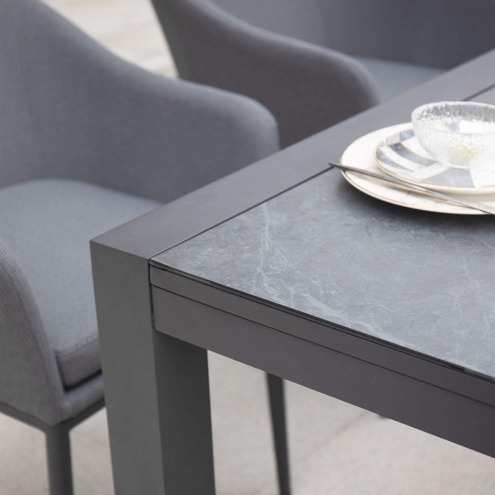 A close-up of the Luna 8 Seat Outdoor Fabric Extending Ceramic Dining Set in Grey, showcasing its modern dining table with a dark, smooth surface and two grey chairs made from outdoor fabric, with plates and bowls placed on the table.
