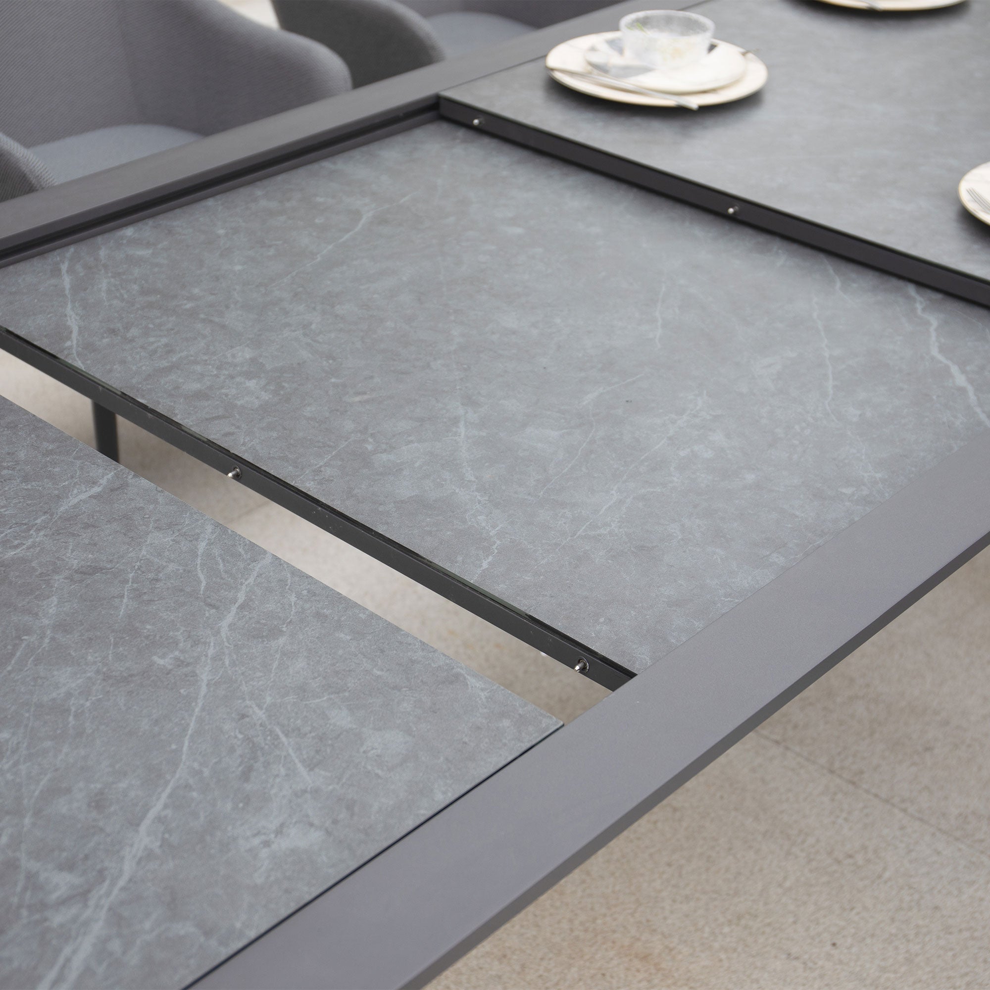 Close-up view of a partially extended Luna 8 Seat Outdoor Fabric Extending Ceramic Dining Set in Grey, with plates and cups on a light-colored floor.