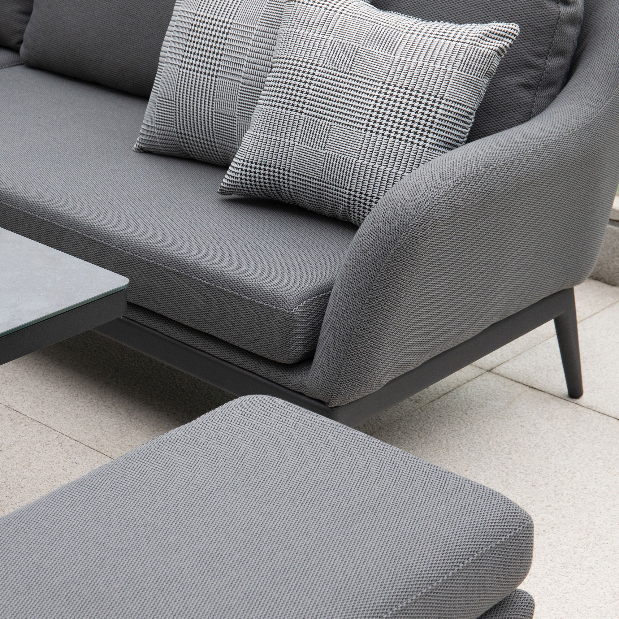 The Luna Deluxe Outdoor Fabric Square Corner Dining Set with Rising Table in Grey includes a sofa with two patterned throw pillows, a matching ottoman, and a gray table on a light stone patio. Crafted from all-weather material, this set promises both style and durability.
