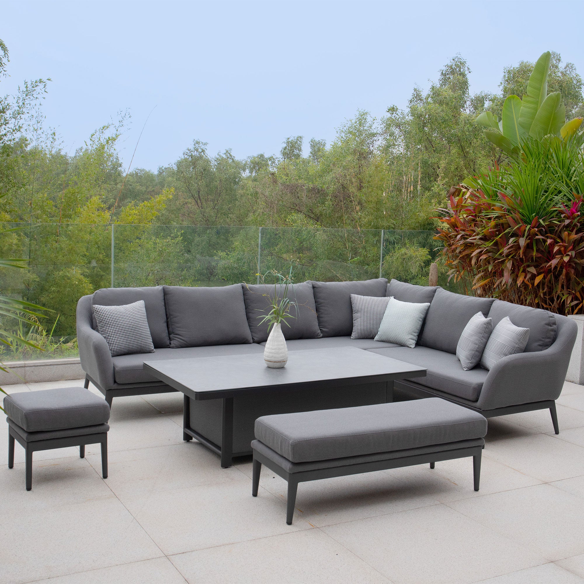 Outdoor patio set featuring the Luna Outdoor Fabric Rectangular Corner Dining Set with Rising Table in Grey (Right Hand), including a sectional sofa, coffee table, ottoman, and bench on a tiled floor with greenery in the background. This Luna outdoor fabric corner dining set adds elegance to any garden space.