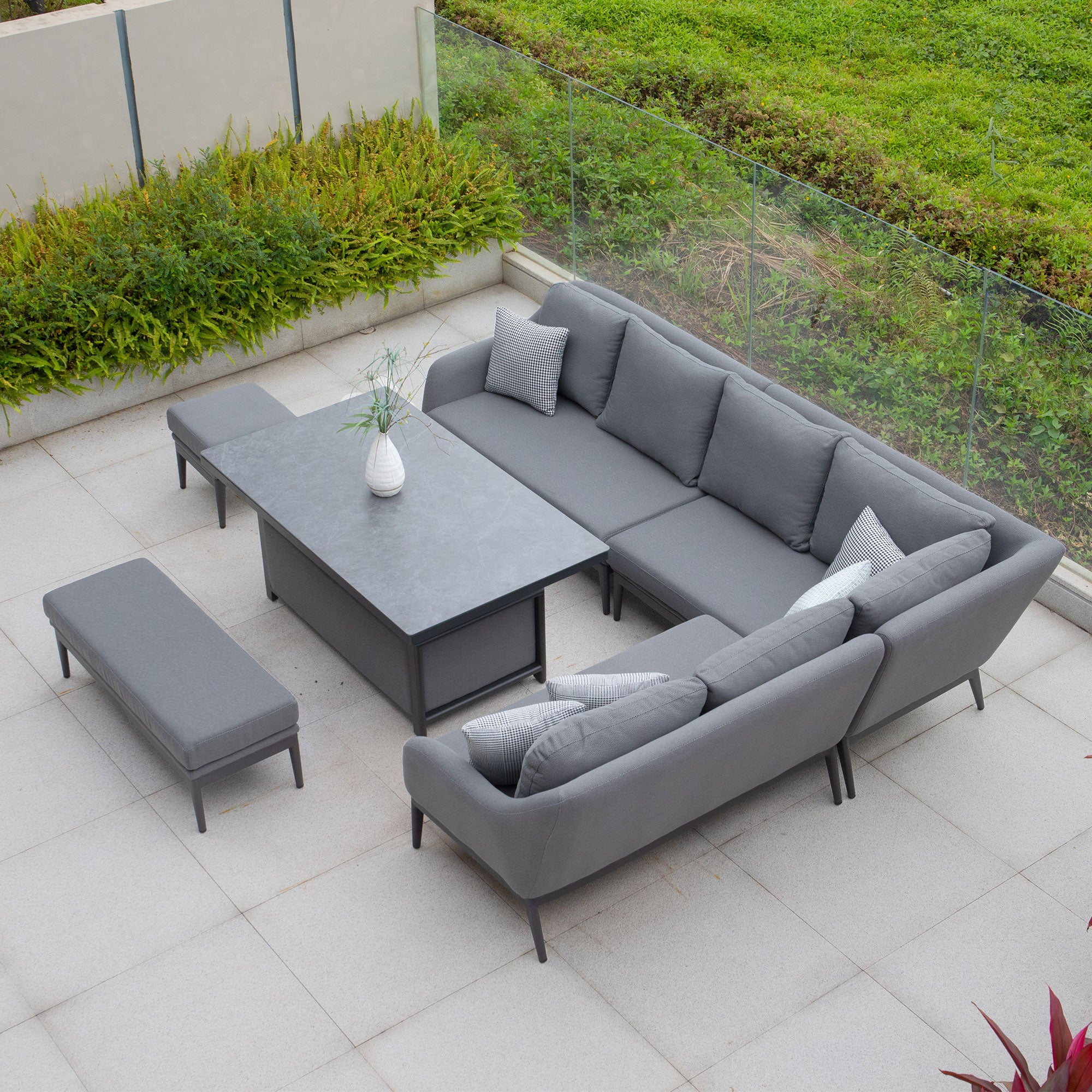 The Luna Outdoor Fabric Rectangular Corner Dining Set with Rising Table in Grey (Right Hand) features a modern gray sectional sofa, matching benches, and a table elegantly positioned on a tiled patio surrounded by lush green foliage. Crafted with superior Luna outdoor fabric, this Harbour Lifestyle set promises both comfort and style for your perfect alfresco experience.