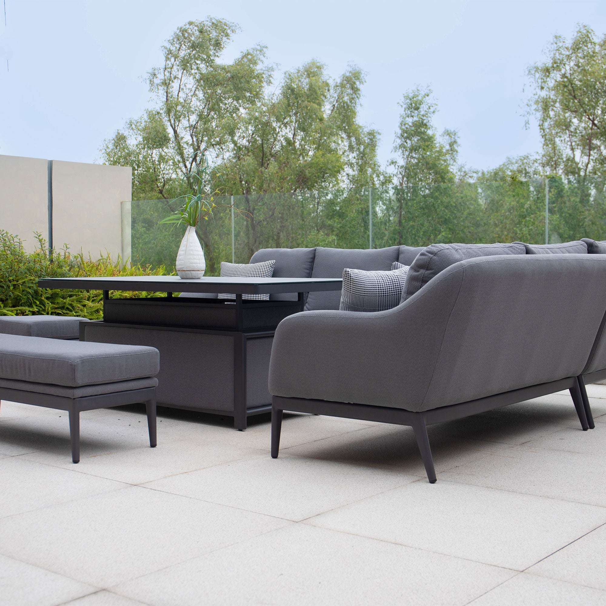 Outdoor patio featuring the Luna Outdoor Fabric Rectangular Corner Dining Set with Rising Table in Grey (Right Hand), accompanied by gray sofas, a matching ottoman, a black table, and green trees in the background.