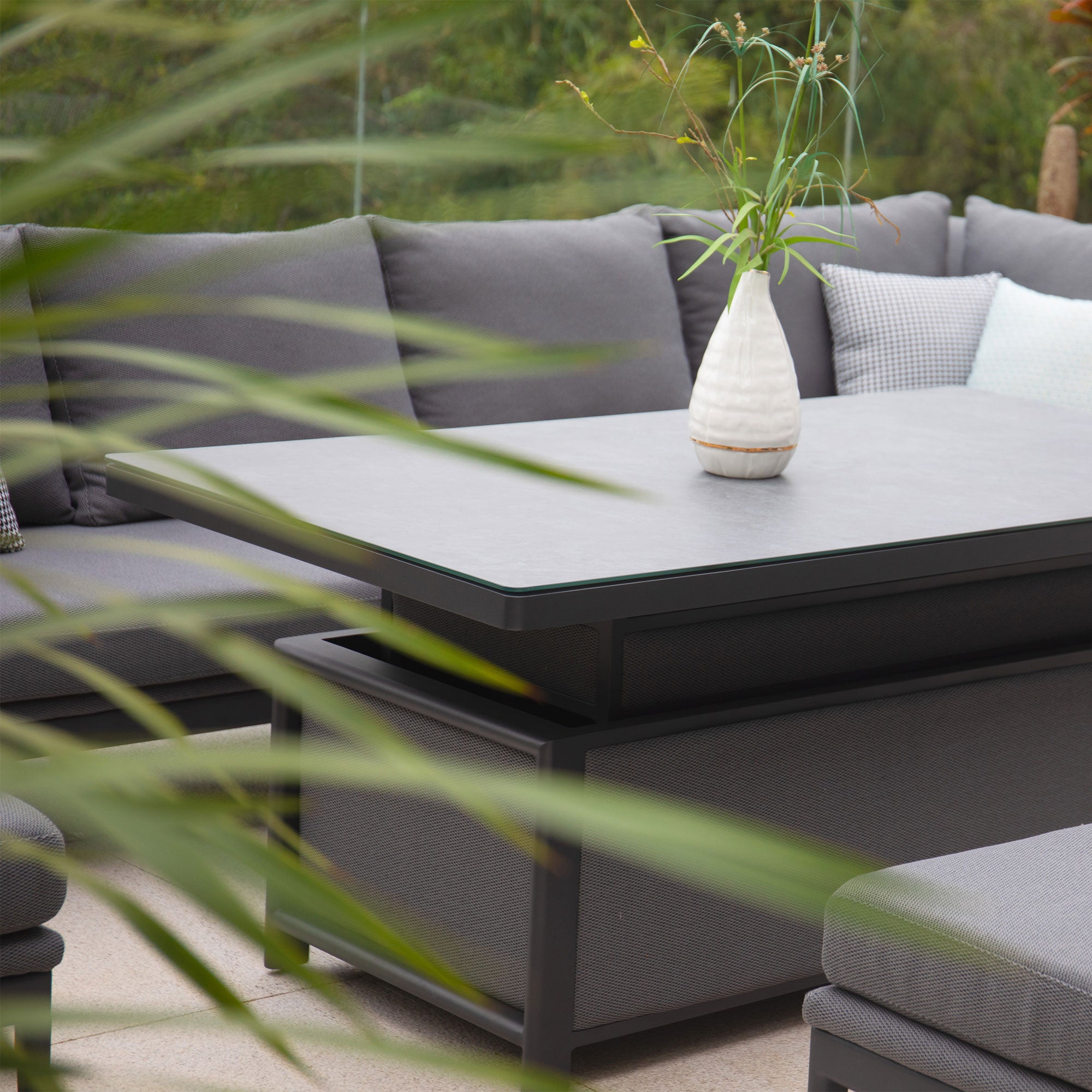 A modern outdoor seating area featuring the Luna Outdoor Fabric Rectangular Corner Dining Set with Rising Table in Grey (Right Hand), complemented by gray cushions, a black table, and a white vase with green plants.