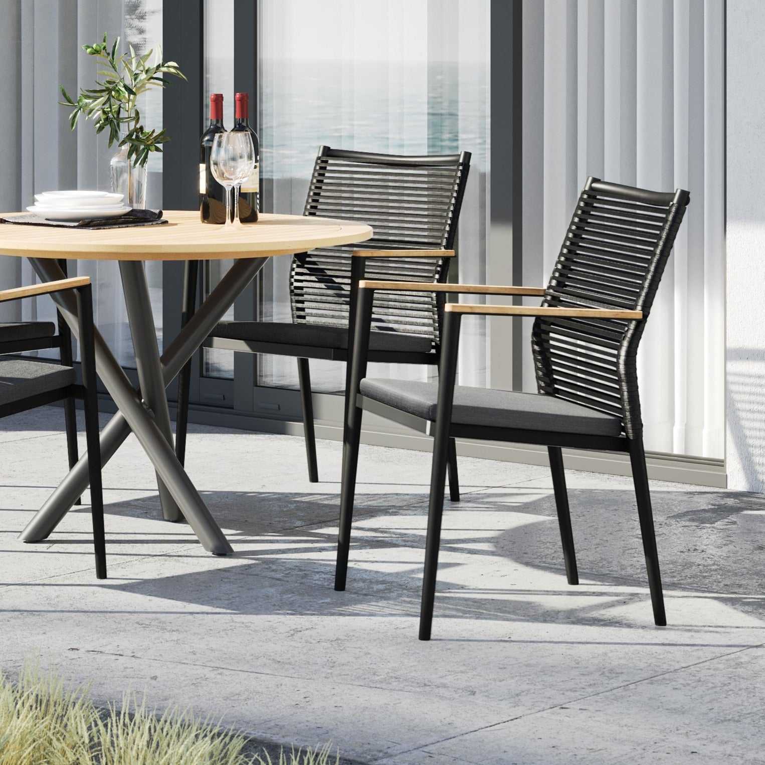 The Portland 4 Seat Round Dining Set with Teak Table in Charcoal includes a teak wood table and two flat rope weaved armchairs with black finishes, complemented by wine bottles and a potted plant.