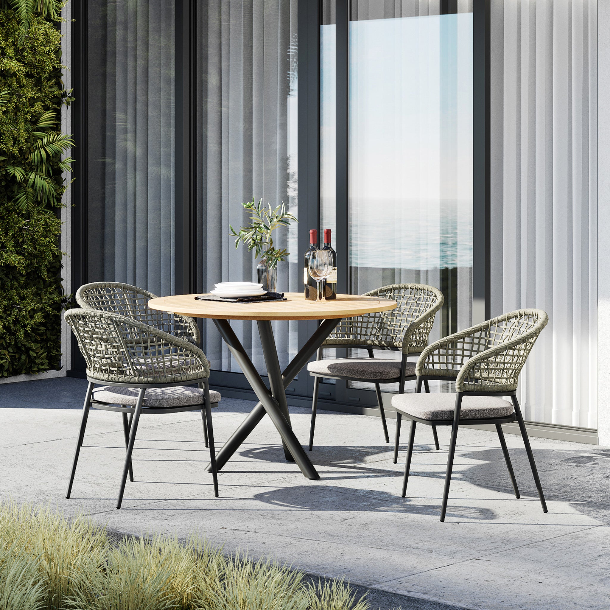 Outdoor patio featuring the Kalama 4 Seat Round Dining Set, which includes a teak table and four woven chairs with sun-proof cushions in olive green, set against the picturesque backdrop of a coastline.