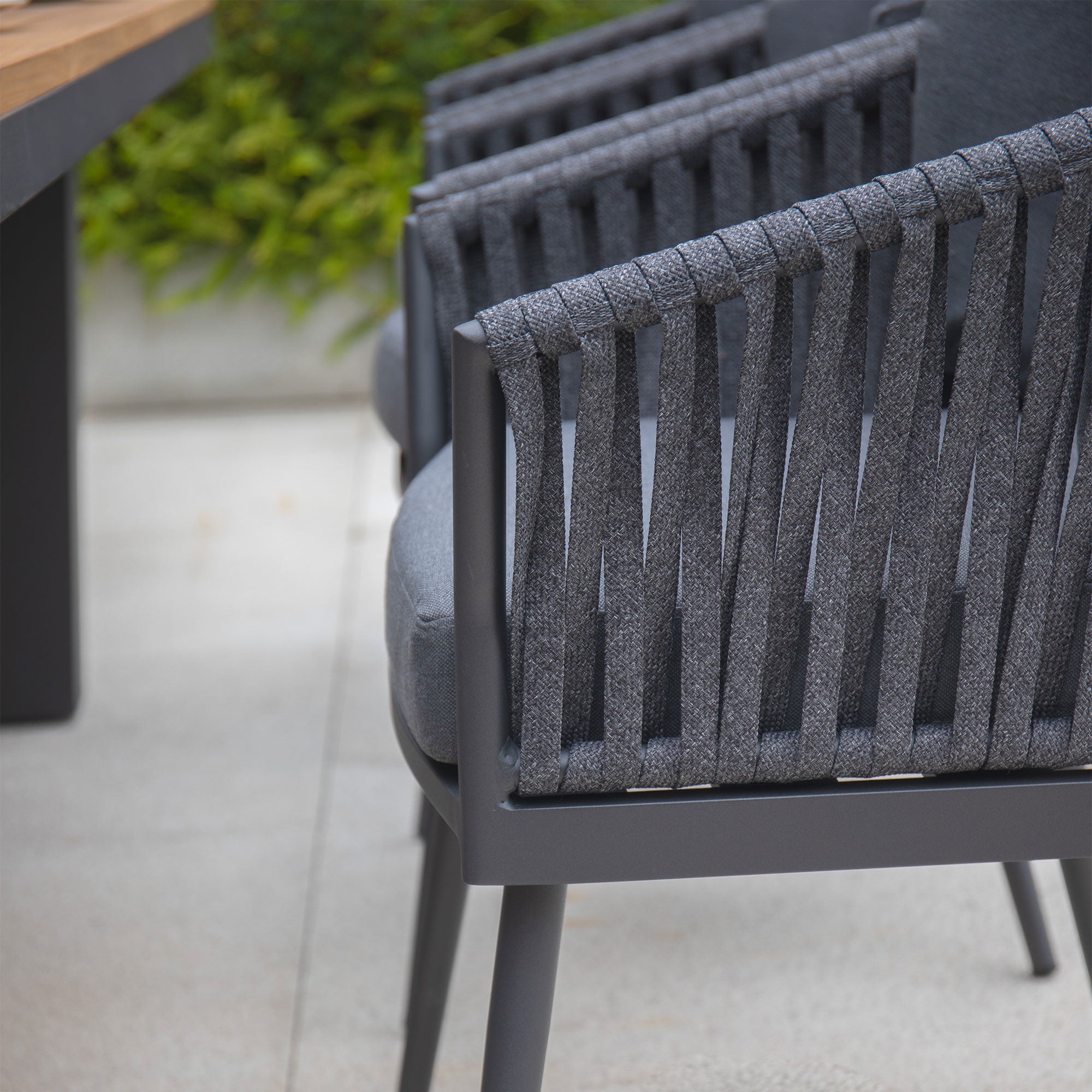 Close-up of the Monterrey 8 Seat Rope Rectangular Teak Firepit Dining Set in Grey from Harbour Lifestyle, featuring gray woven-back outdoor chairs next to a wooden table on a patio with lush greenery in the background.
