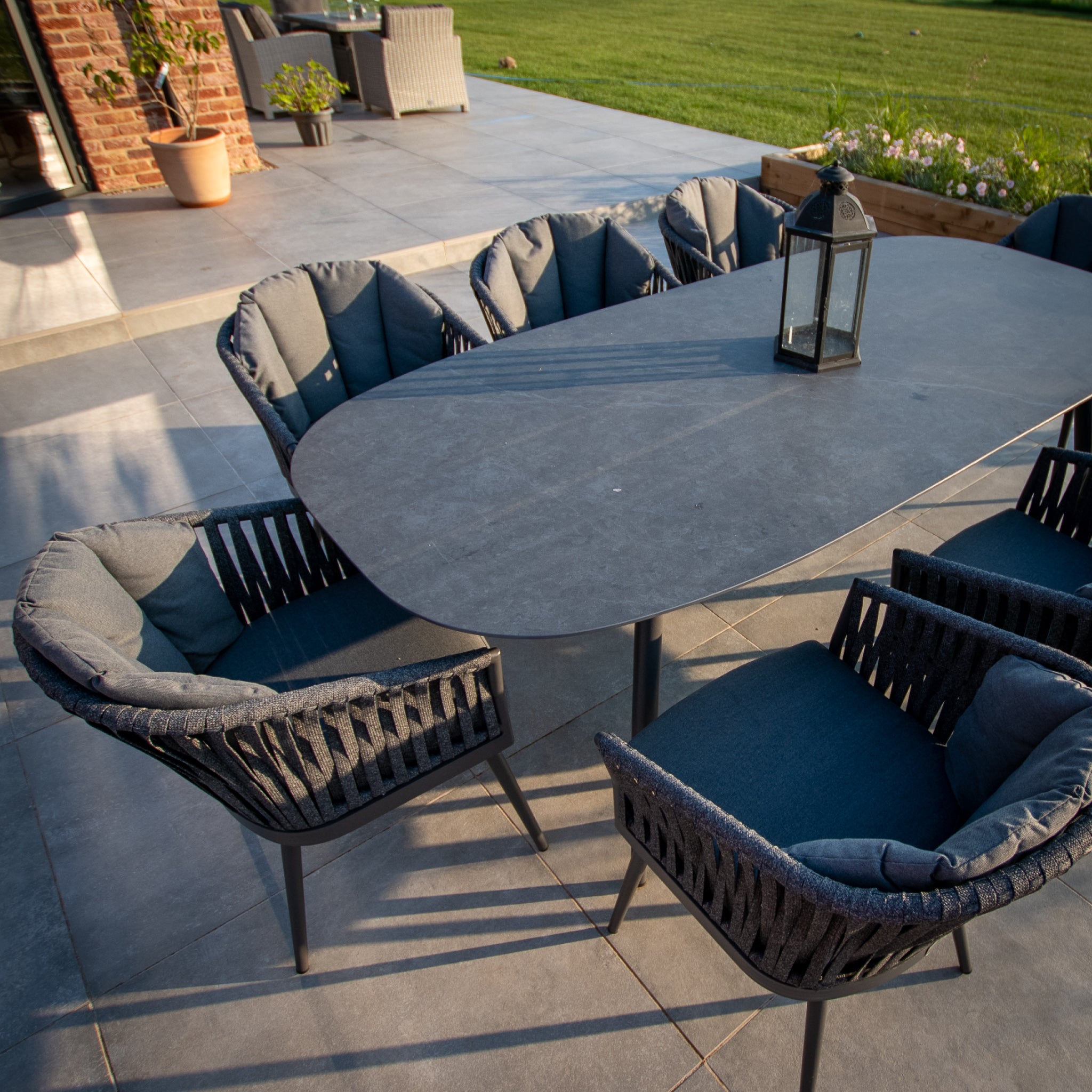 The Monterrey 8 Seat Rope Dining Set with an oval ceramic table in grey features cushioned chairs and a black lantern centerpiece, all beautifully arranged on a tiled terrace from Harbour Lifestyle.