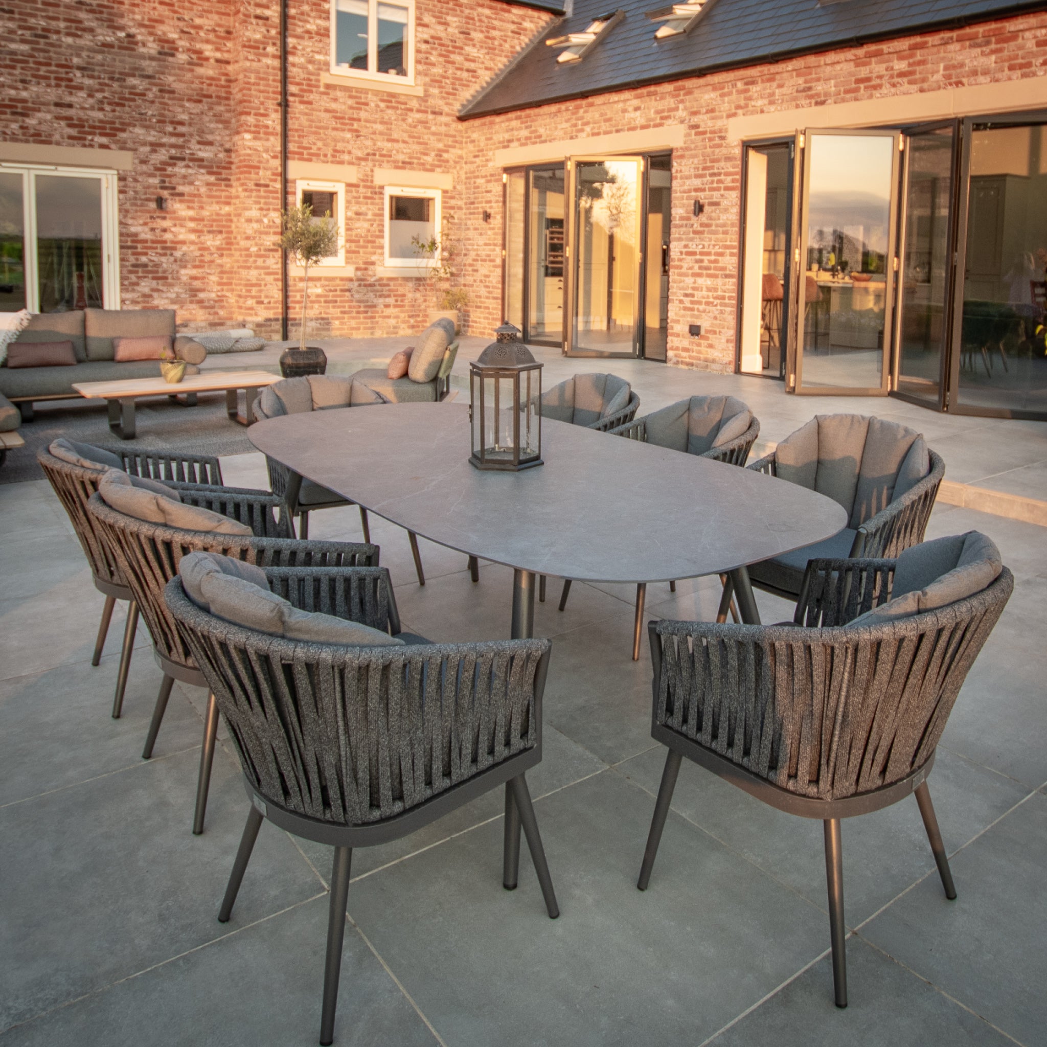 Outdoor patio featuring the Monterrey 8 Seat Rope Dining Set with an oval ceramic table in grey, complemented by a lantern centerpiece and set against a modern brick house backdrop.