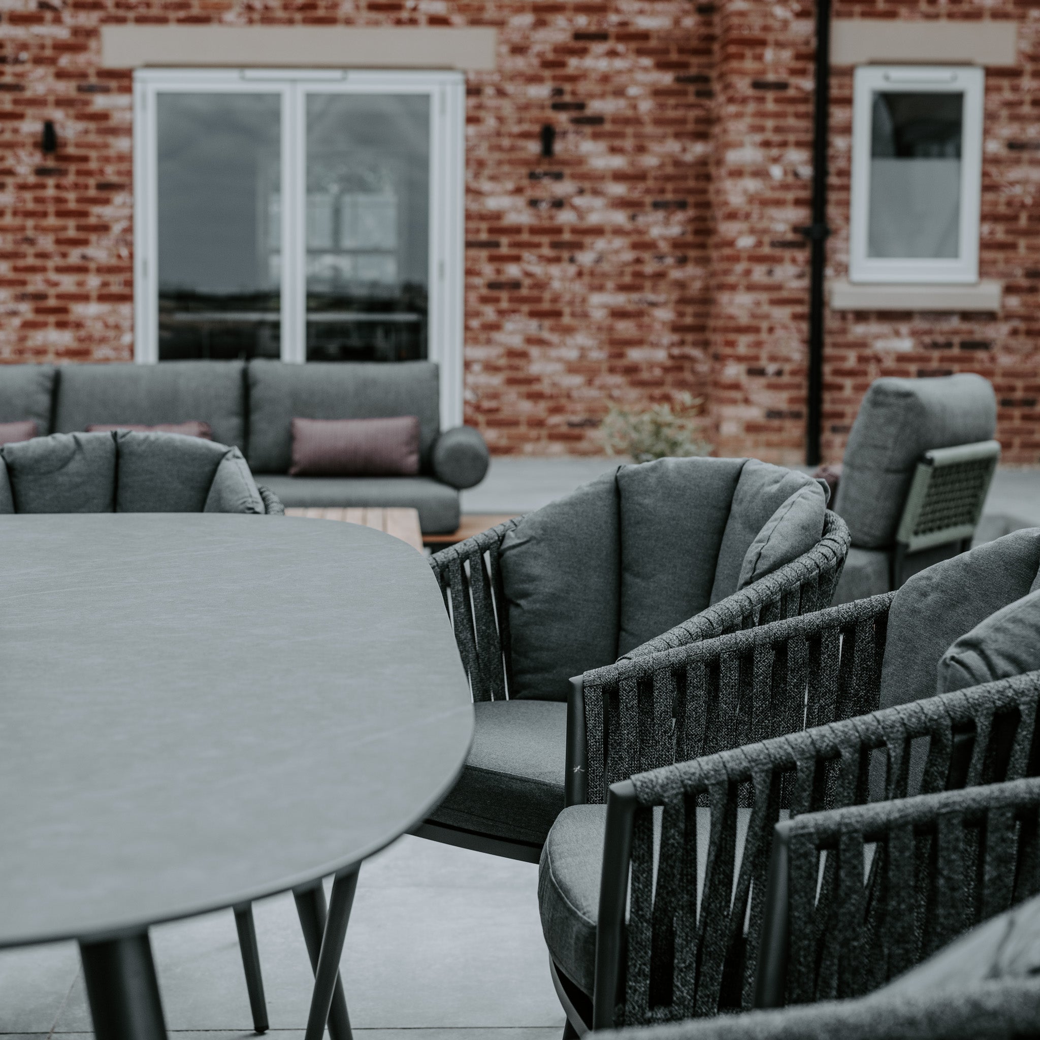 Outdoor patio featuring the Monterrey 8 Seat Rope Dining Set with an Oval Ceramic Table in Grey, cushioned chairs, all set against a backdrop of a brick wall with large windows.