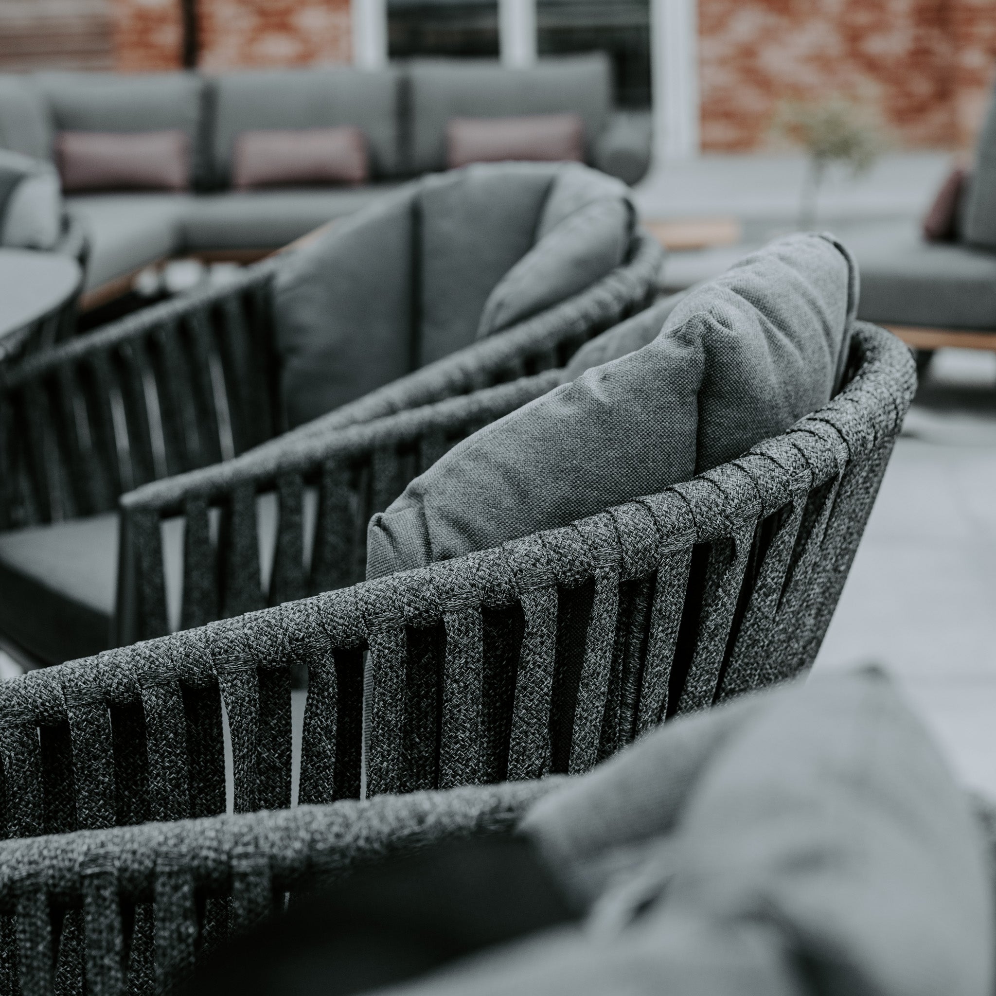 Close-up of a stylish outdoor seating area featuring gray cushioned chairs and a sofa in the background on a patio, paired perfectly with the Monterrey 8 Seat Rope Dining Set with Oval Ceramic Table in Grey from Harbour Lifestyle.