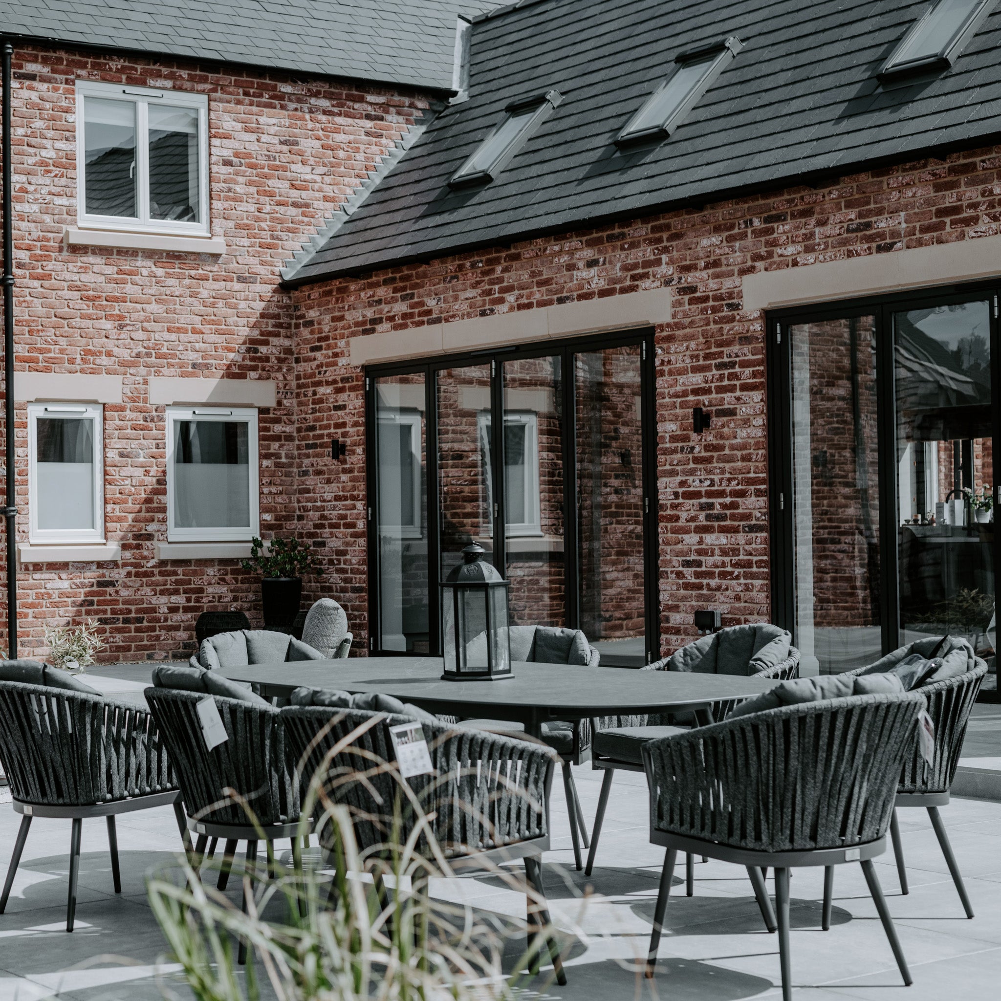 Modern patio featuring a Monterrey 8 Seat Rope Dining Set with Oval Ceramic Table in Grey, set against a red brick house with large sliding glass doors and skylights.