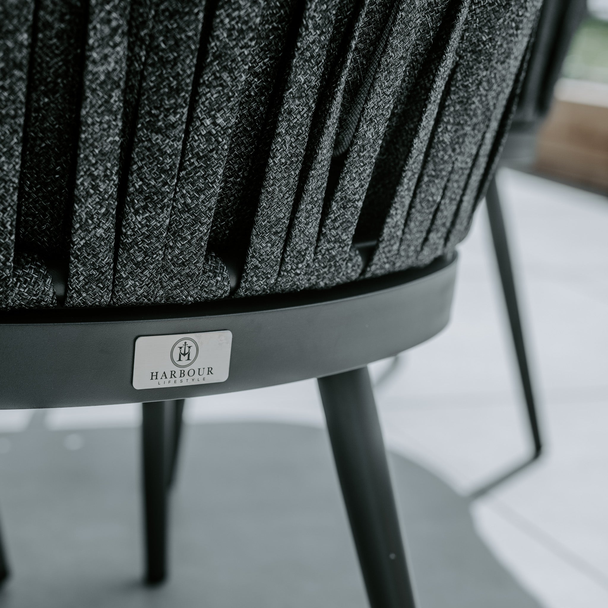 Close-up of a stylish, fabric-covered chair with a metal base, featuring a "HARBOUR" label on the backrest, complemented by the sophisticated elegance of the Monterrey 8 Seat Rope Dining Set with Oval Ceramic Table in Grey.